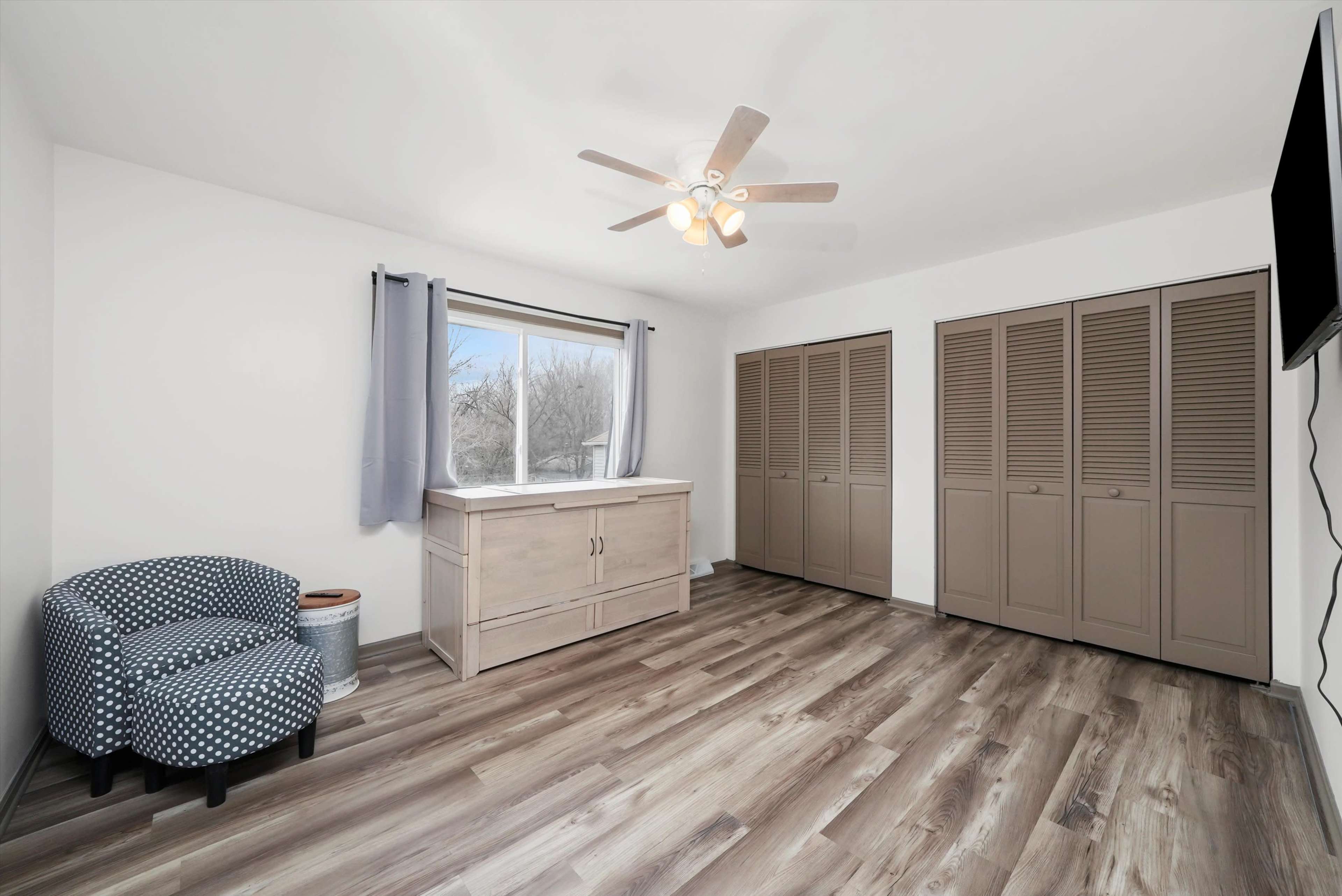 A spacious bedroom featuring a ceiling fan, a window with curtains, a wooden chest, a patterned chair, and multiple storage closets with louvered doors.