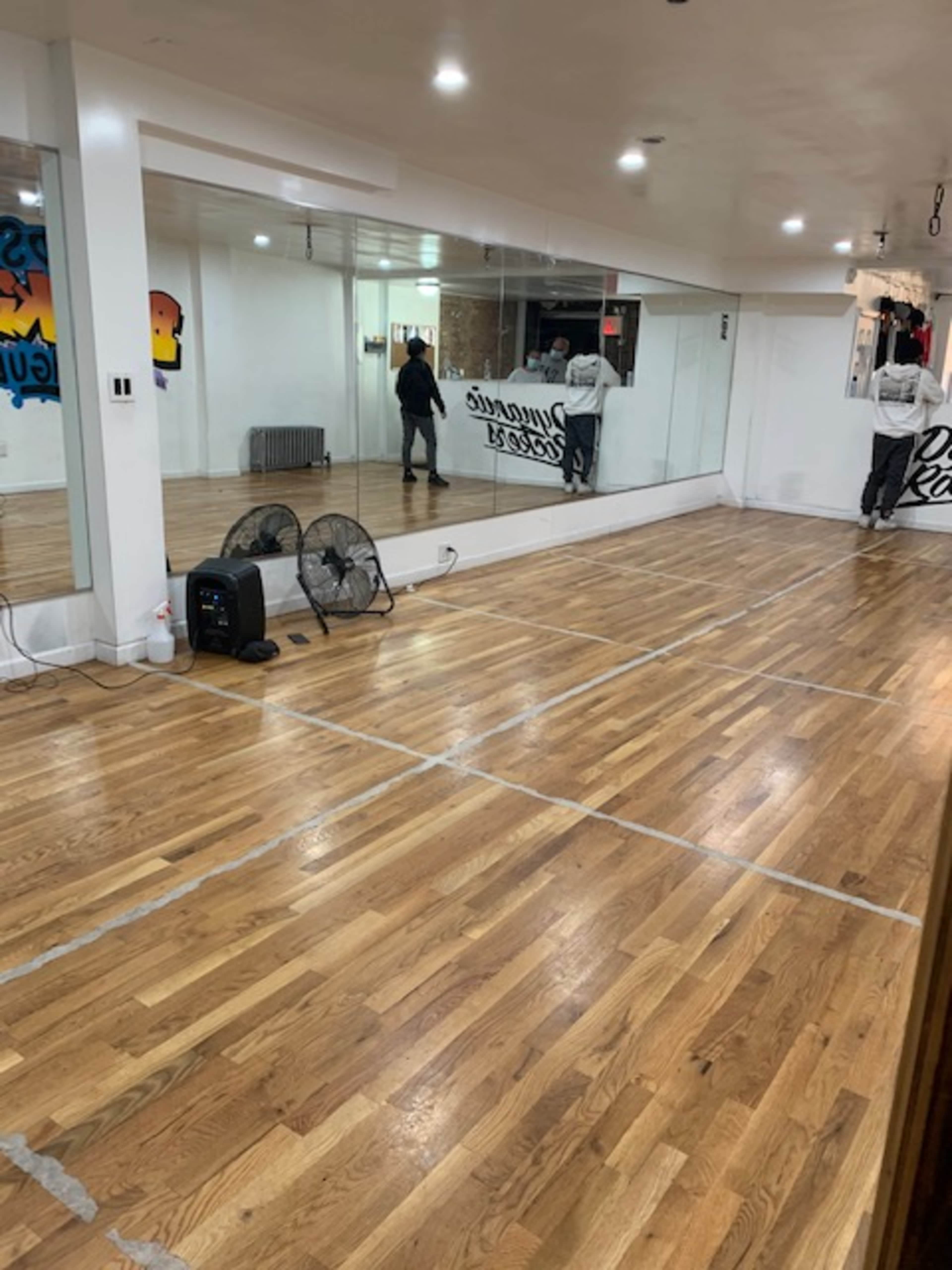 A spacious dance studio with polished wooden flooring, mirrors on one wall, and two standing fans along the side.