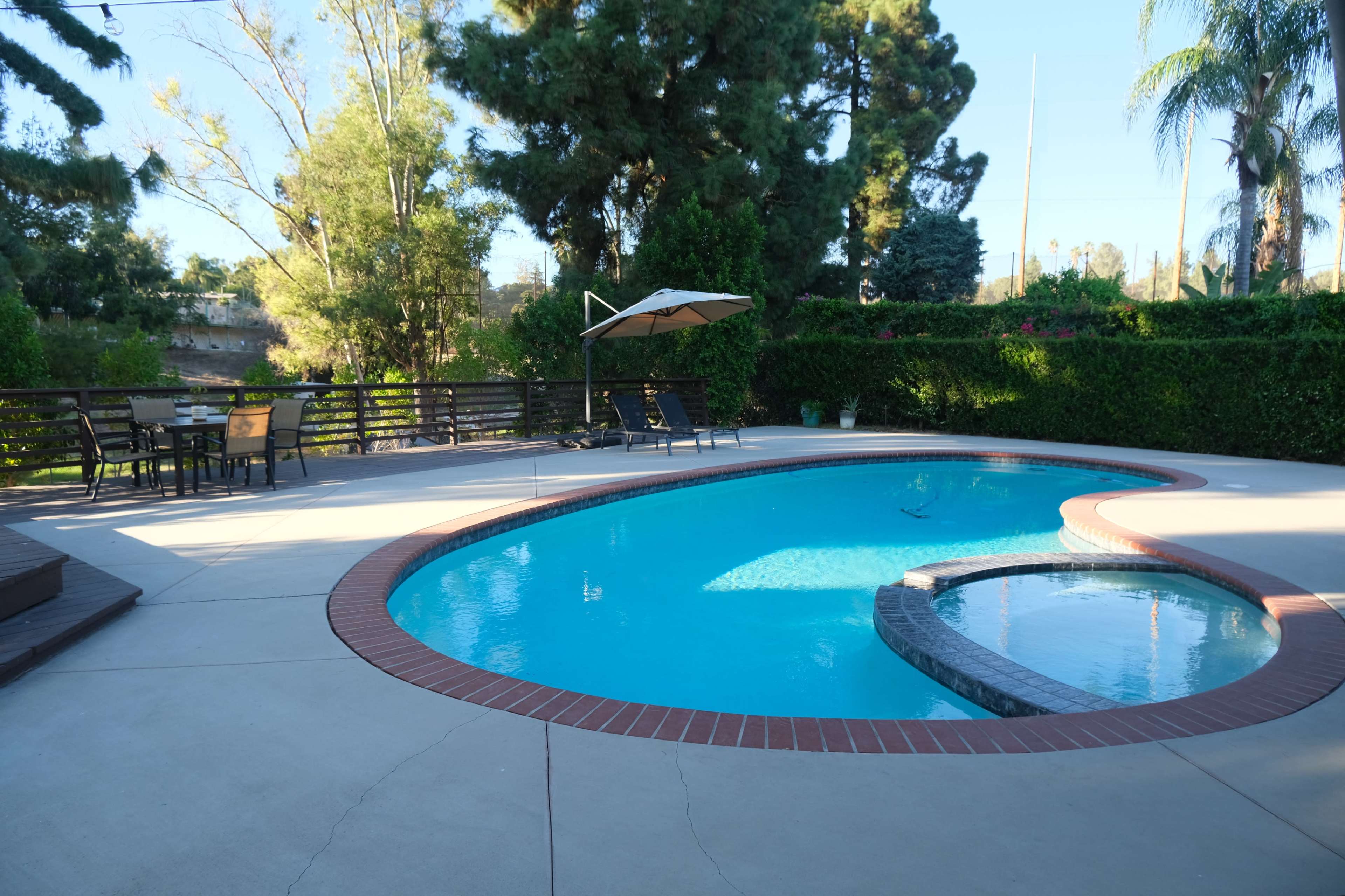 The image shows a backyard with a swimming pool, surrounded by trees and a seating area featuring a table and chairs.
