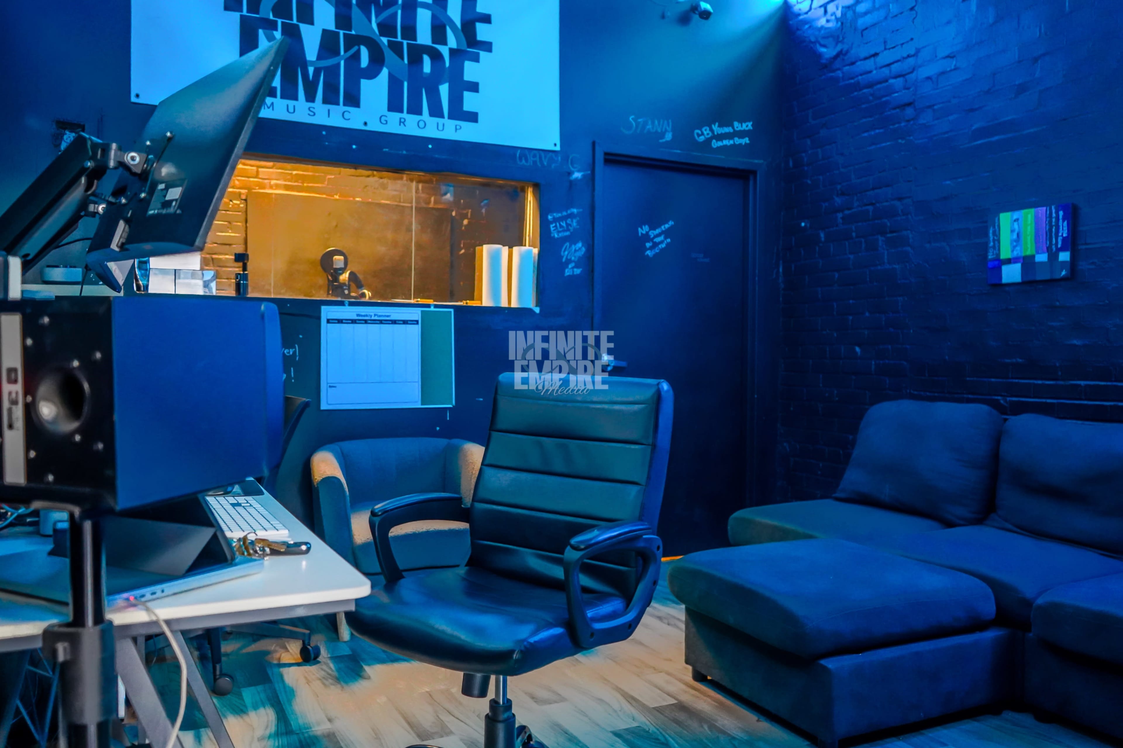 The image depicts a cozy music studio with a black ergonomic chair in front of a computer desk and a large dark couch in the corner.