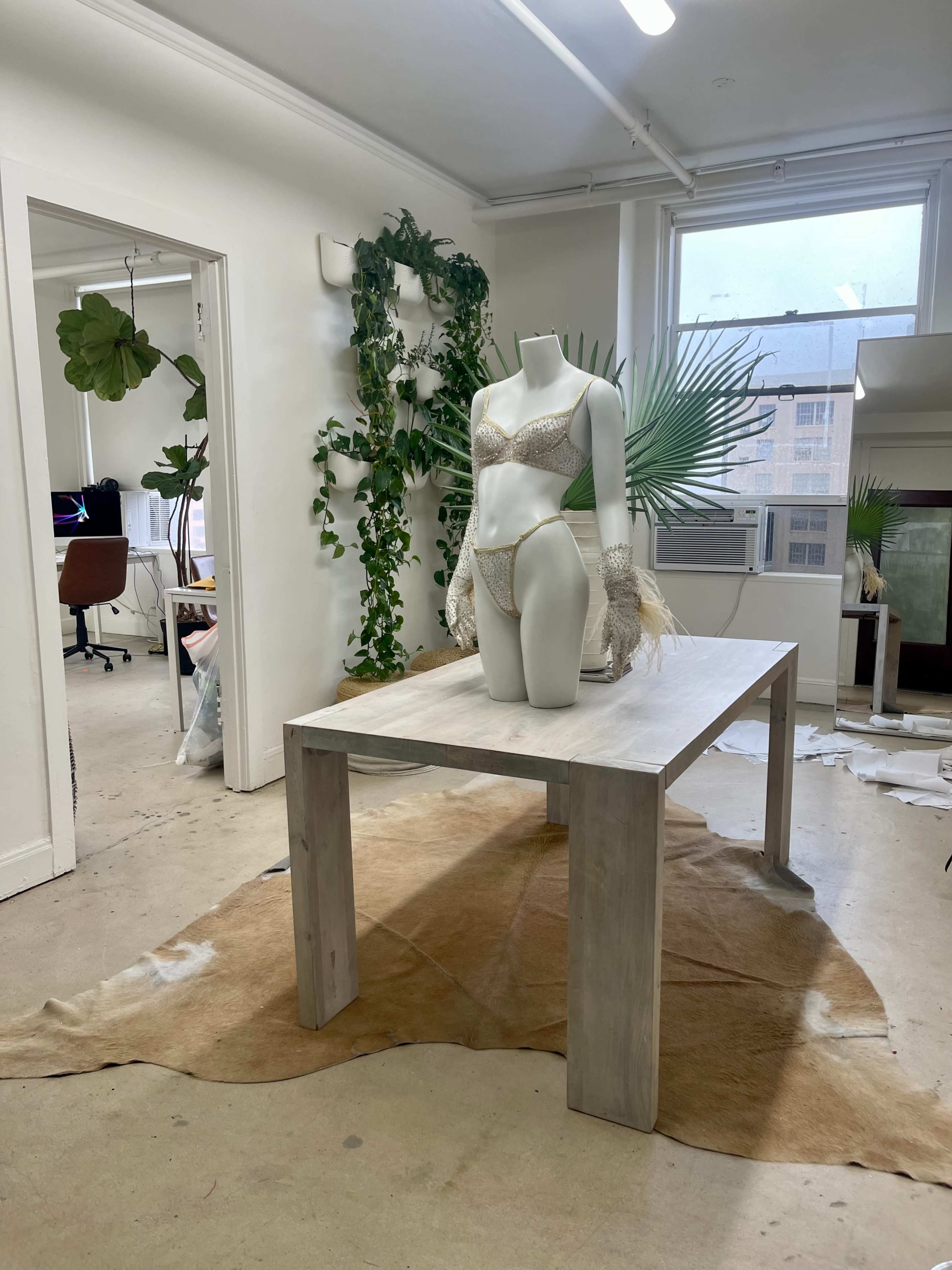 A white mannequin in a bikini stands on a wooden table surrounded by plants in a bright, open workspace.