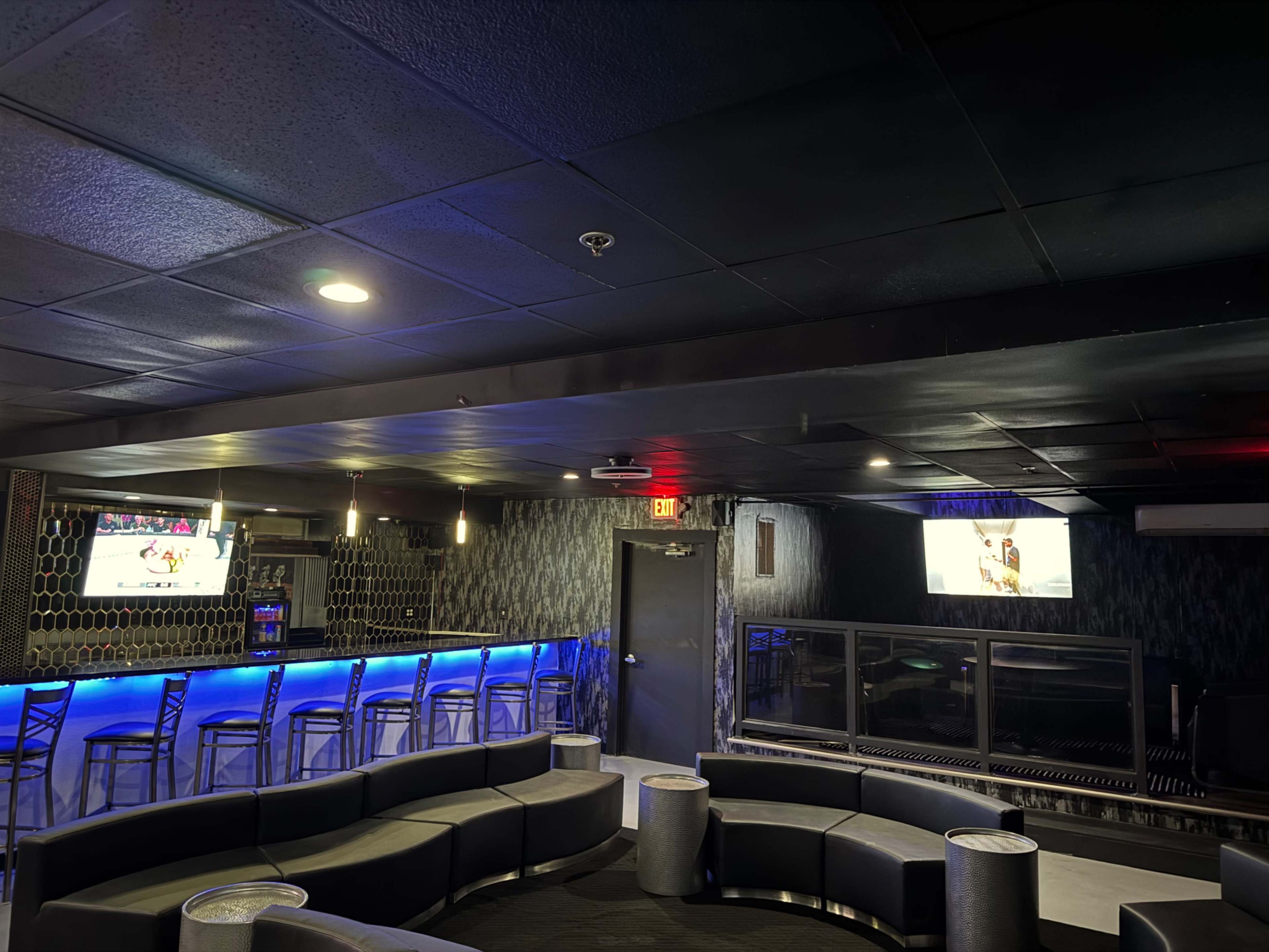 The image shows a dimly lit lounge area with a curved seating arrangement, a bar with high stools, and two screens showing content on the walls.
