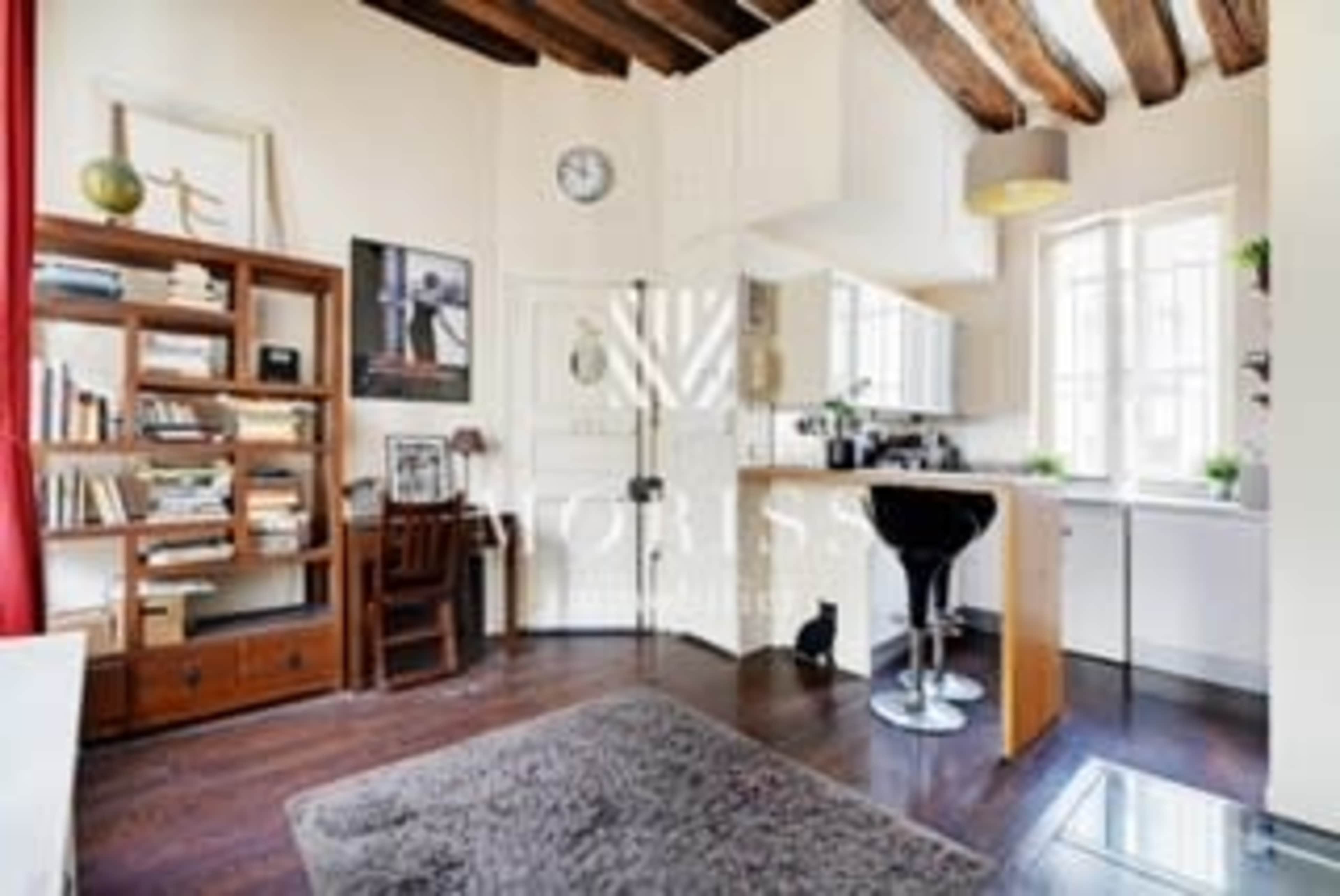 The image shows a small, well-lit room with wooden beams, a bookshelf filled with books, a kitchenette, and a bar-style seating area.
