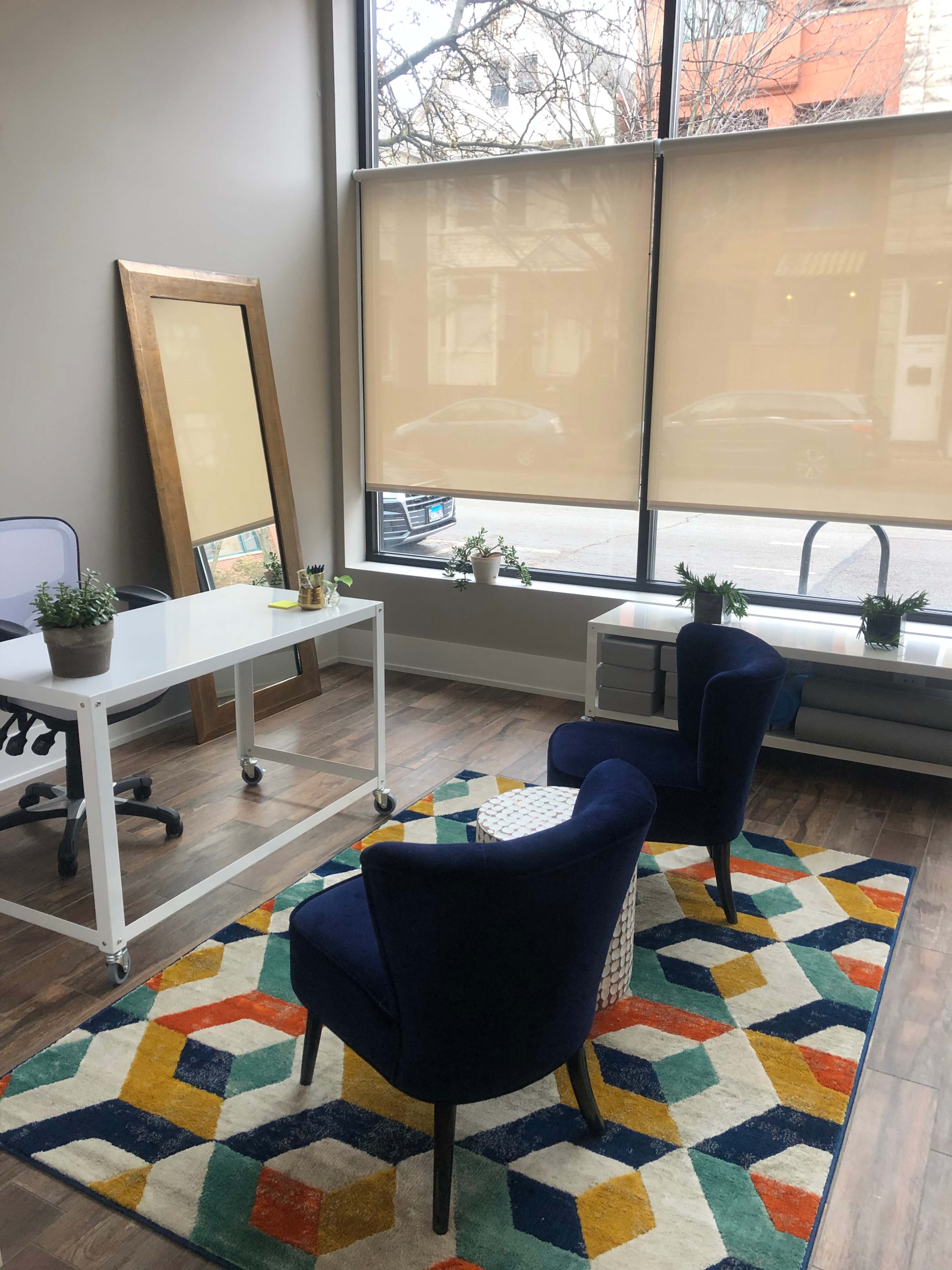 A workspace with two blue chairs, a white desk, and large windows covered by shades, revealing a city street outside.