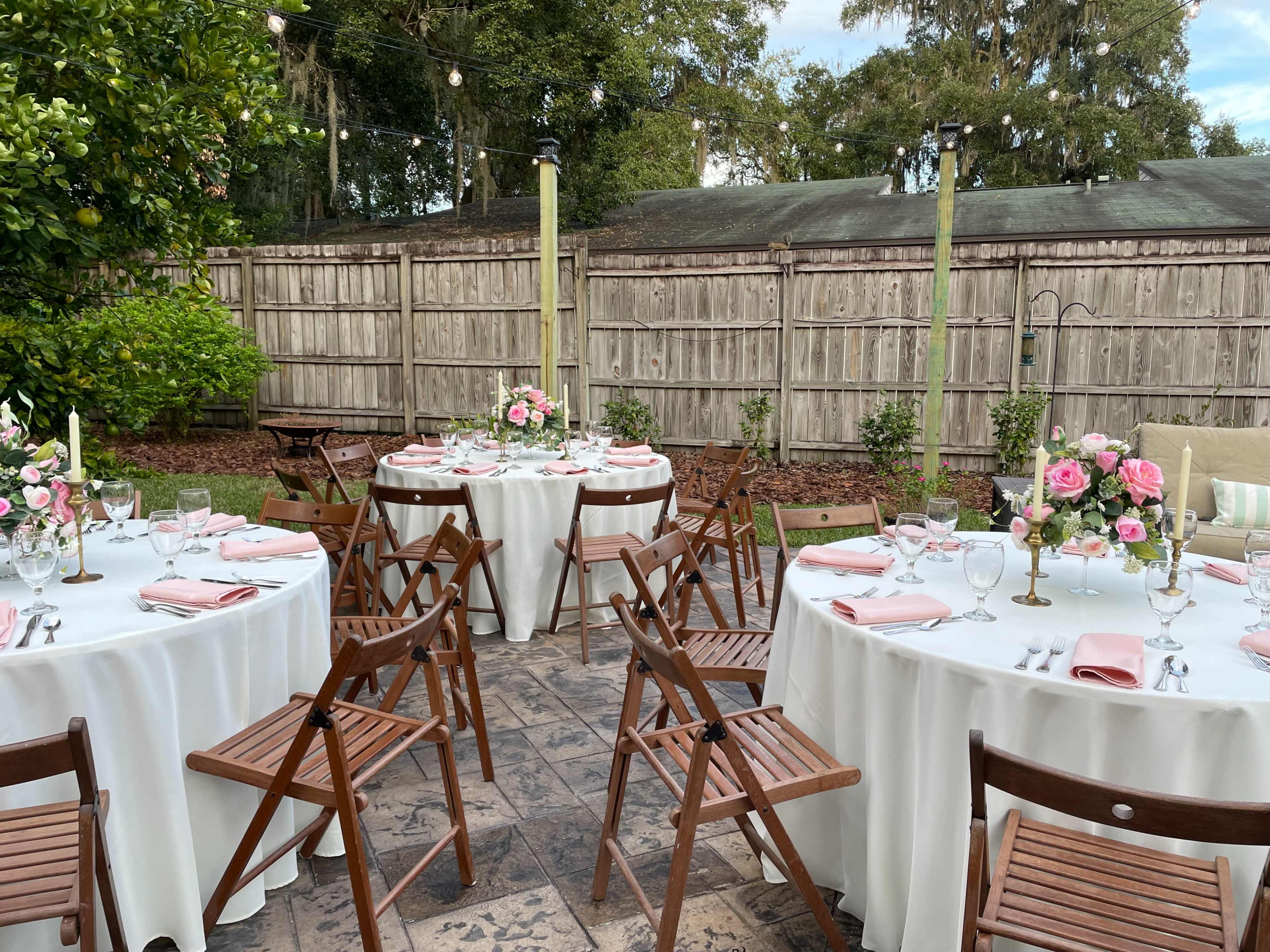 The image shows a decorated outdoor dining area with round tables covered in white tablecloths and set with pink napkins, surrounded by wooden chairs and floral centerpieces.