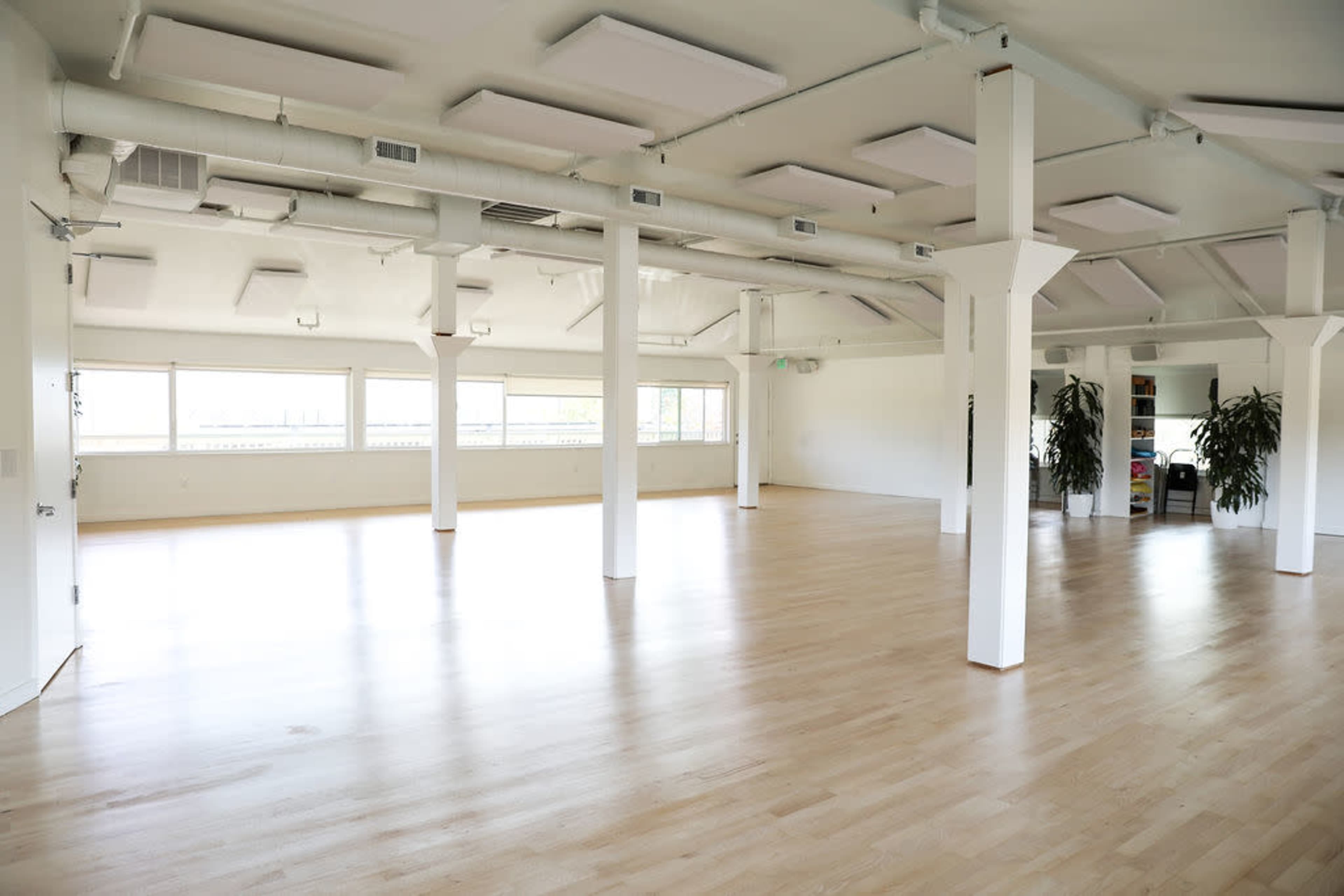 A spacious, empty room with wooden flooring, large windows, and white walls, supported by tall columns.