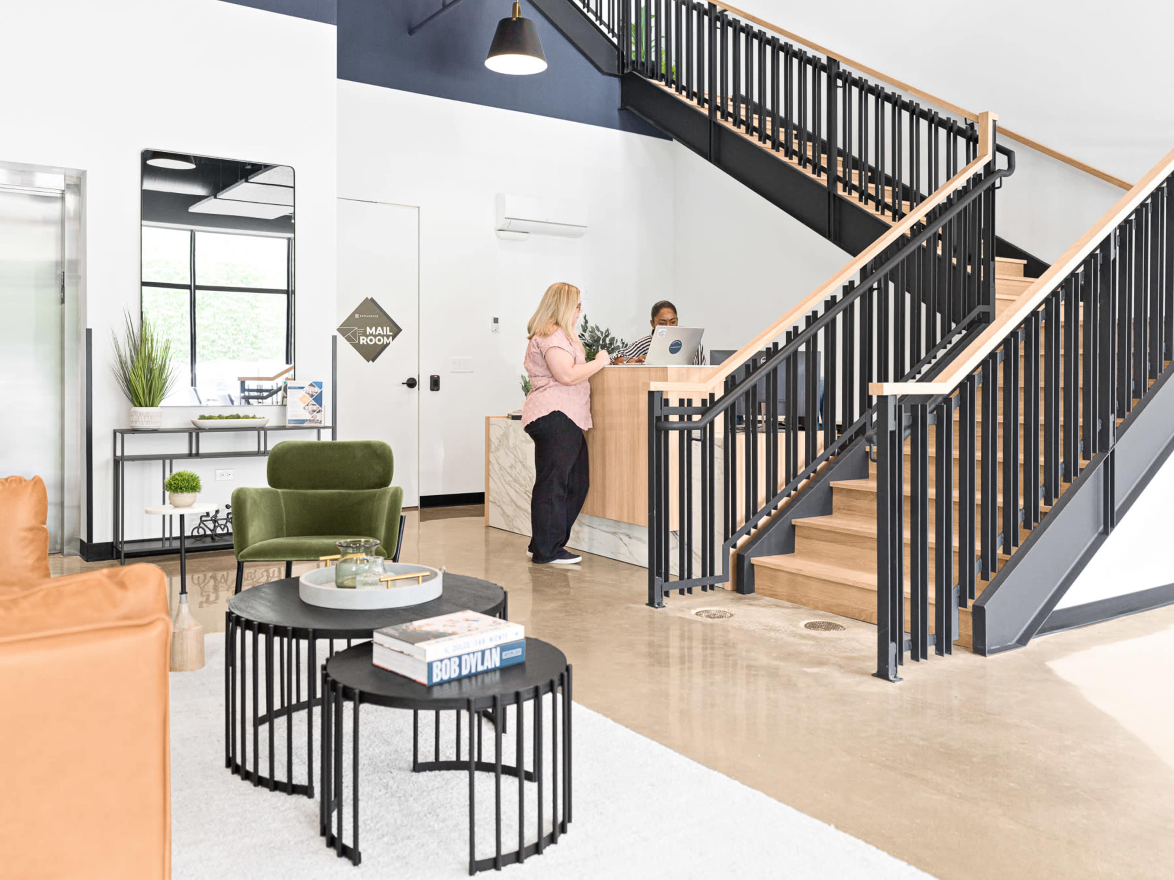 The image shows a modern reception area with a staircase, a seating area featuring green chairs, and a reception desk where a staff member is assisting a visitor.
