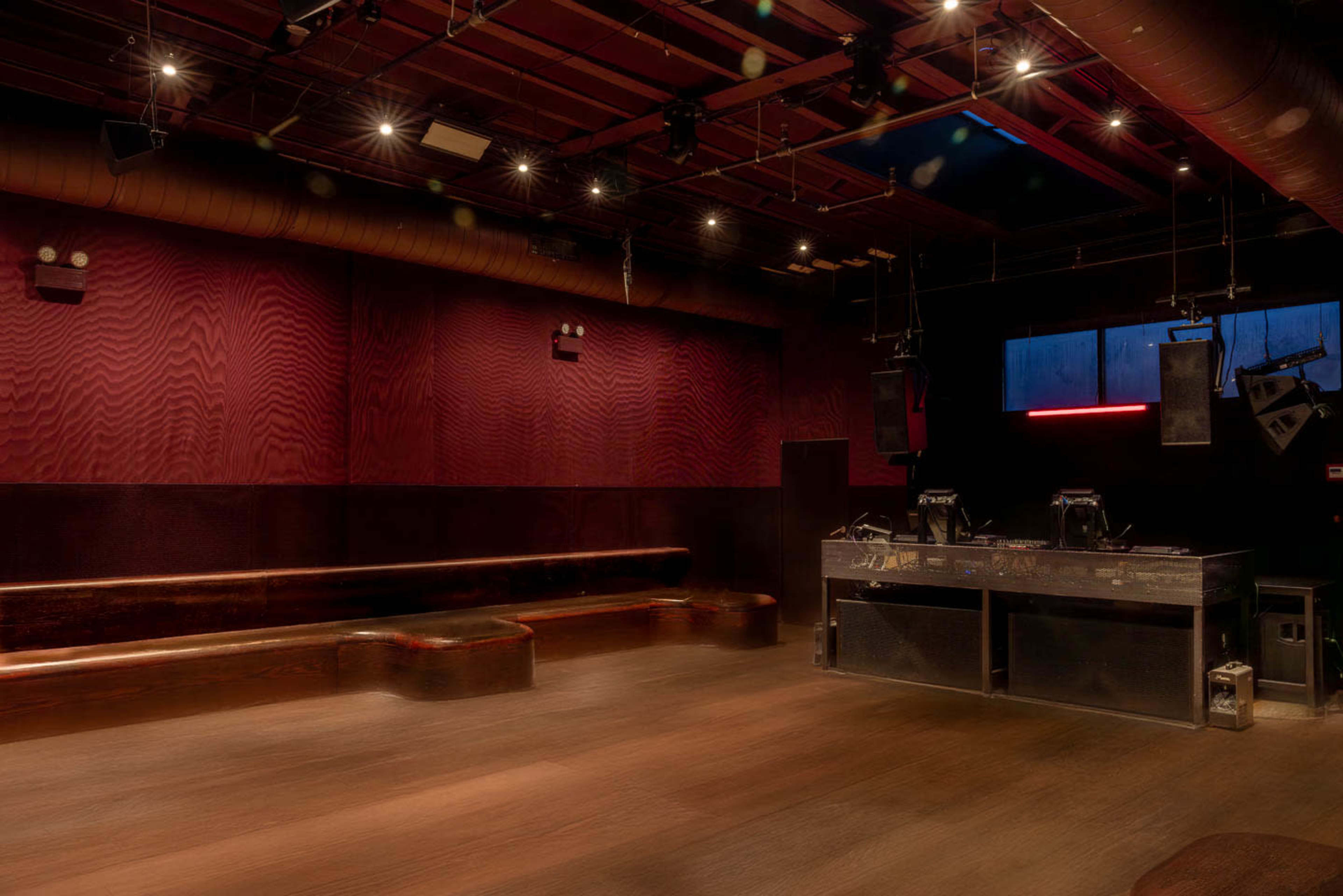 The image shows a dimly lit indoor venue with dark walls, wooden flooring, and a bar area featuring sound equipment.