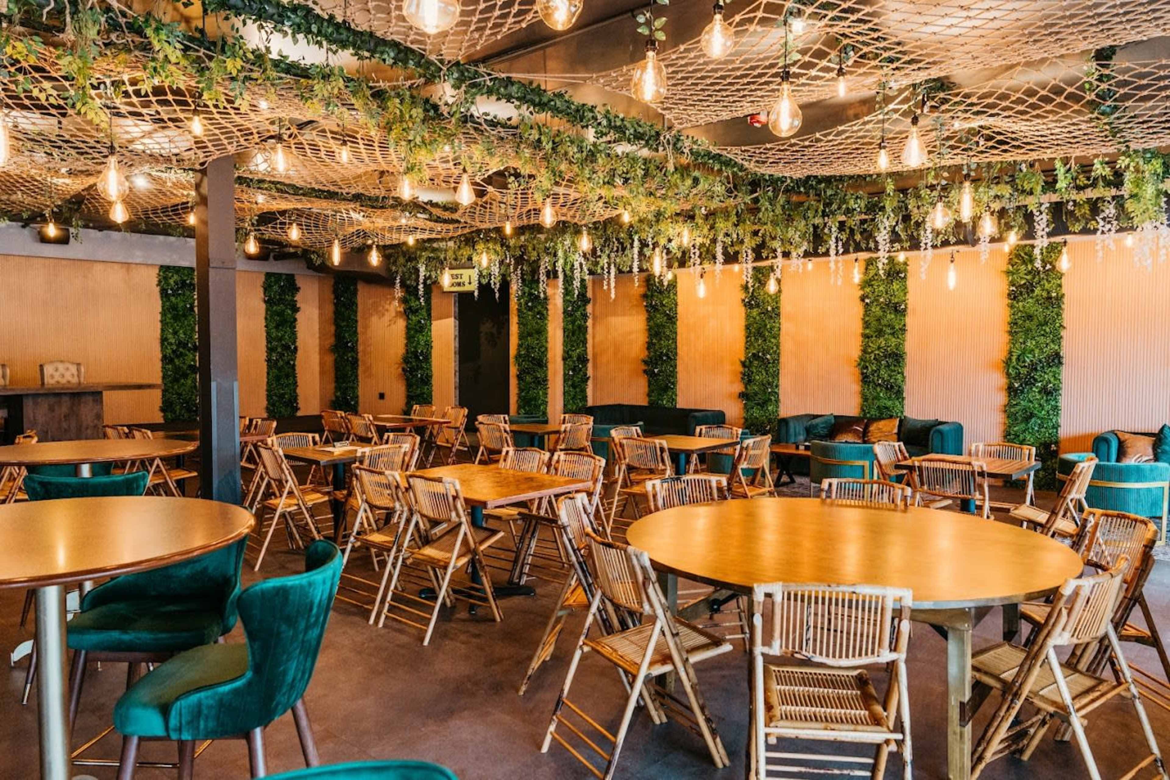 A decorated indoor venue features wooden tables and chairs surrounded by greenery and hanging lighting.