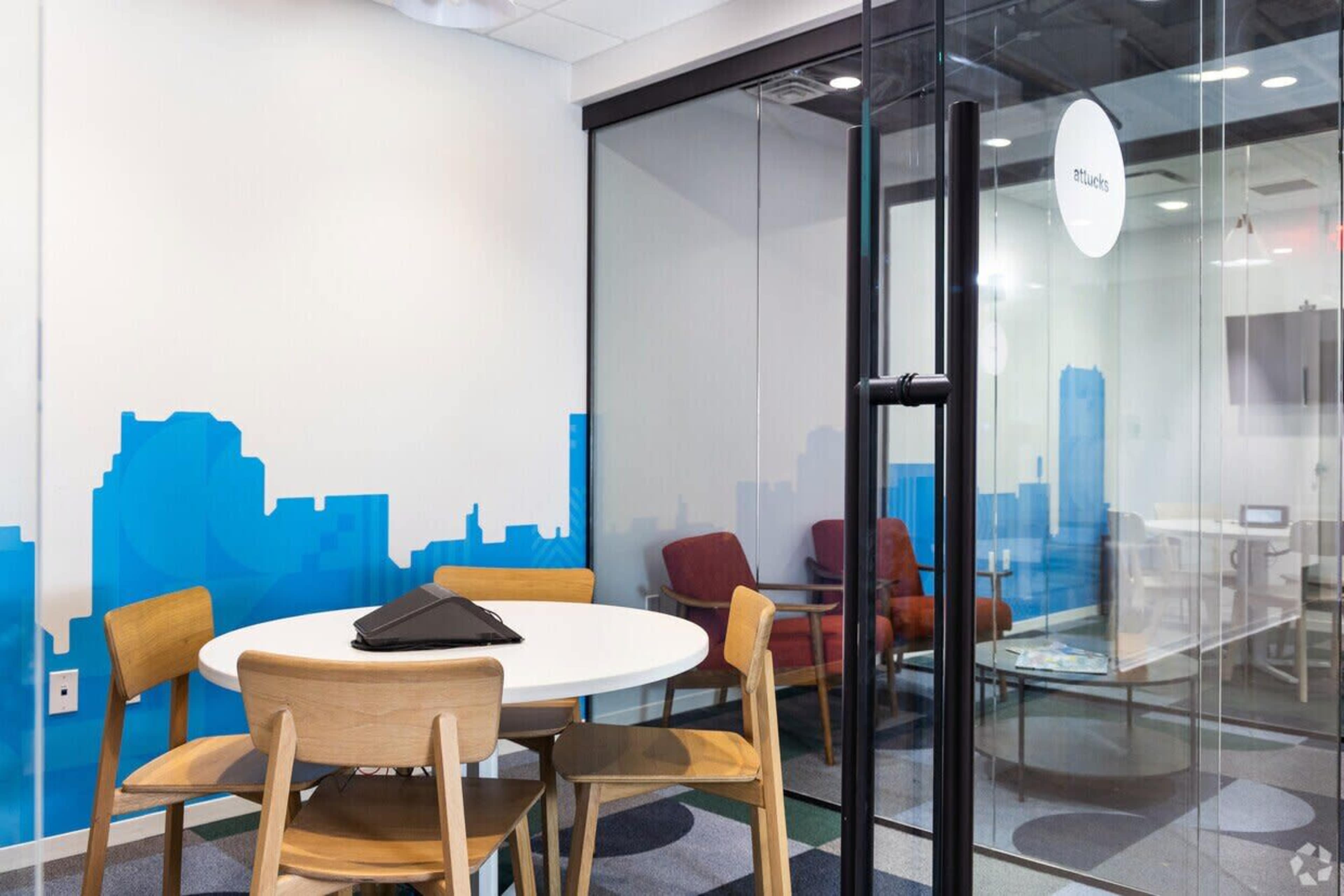 A bright meeting room features a round table surrounded by wooden chairs, with a glass wall displaying a city skyline graphic in the background.