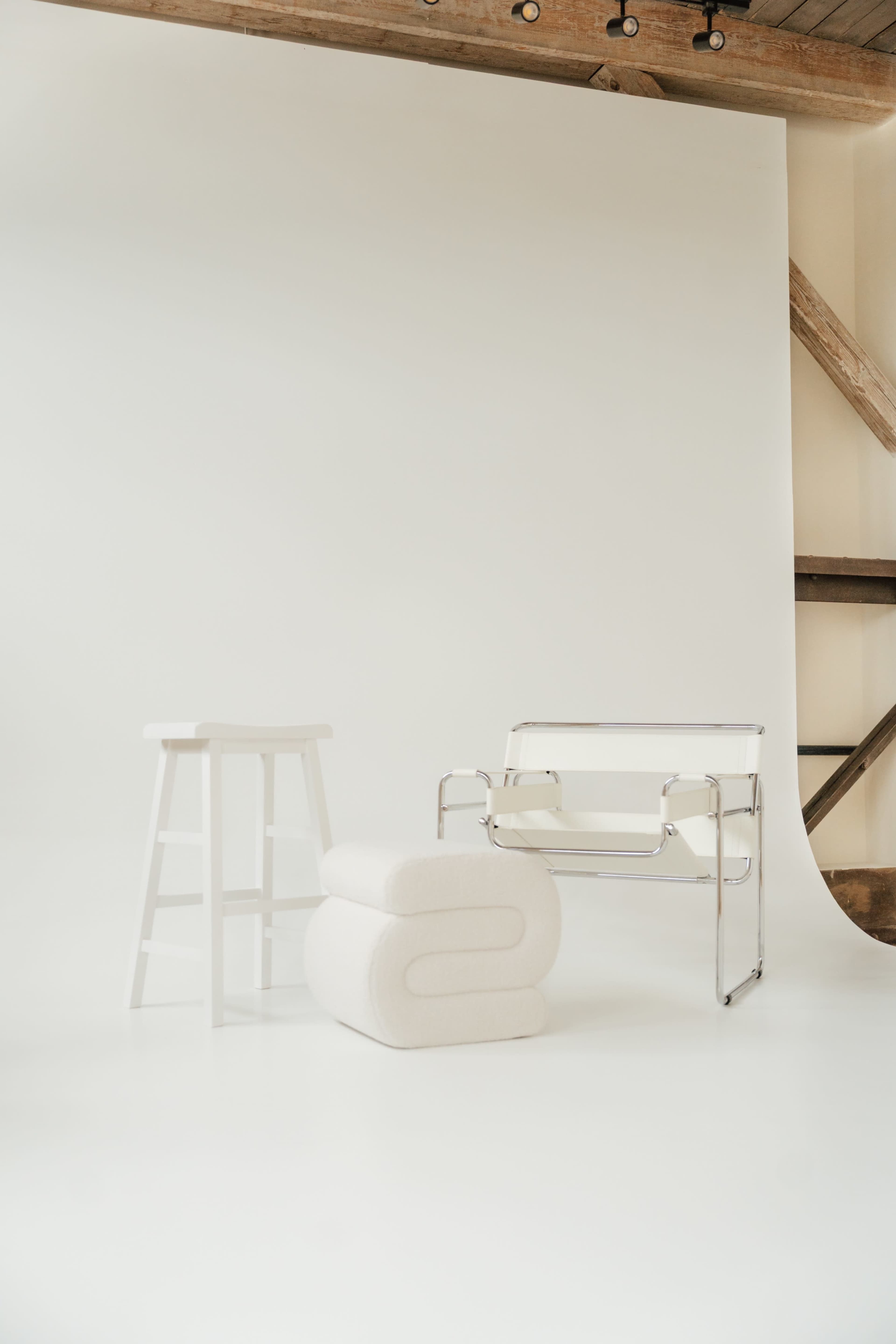 The image features a minimalist interior scene with a white background, showcasing a chrome-framed chair, a white stool, and a two-piece padded seating arrangement.