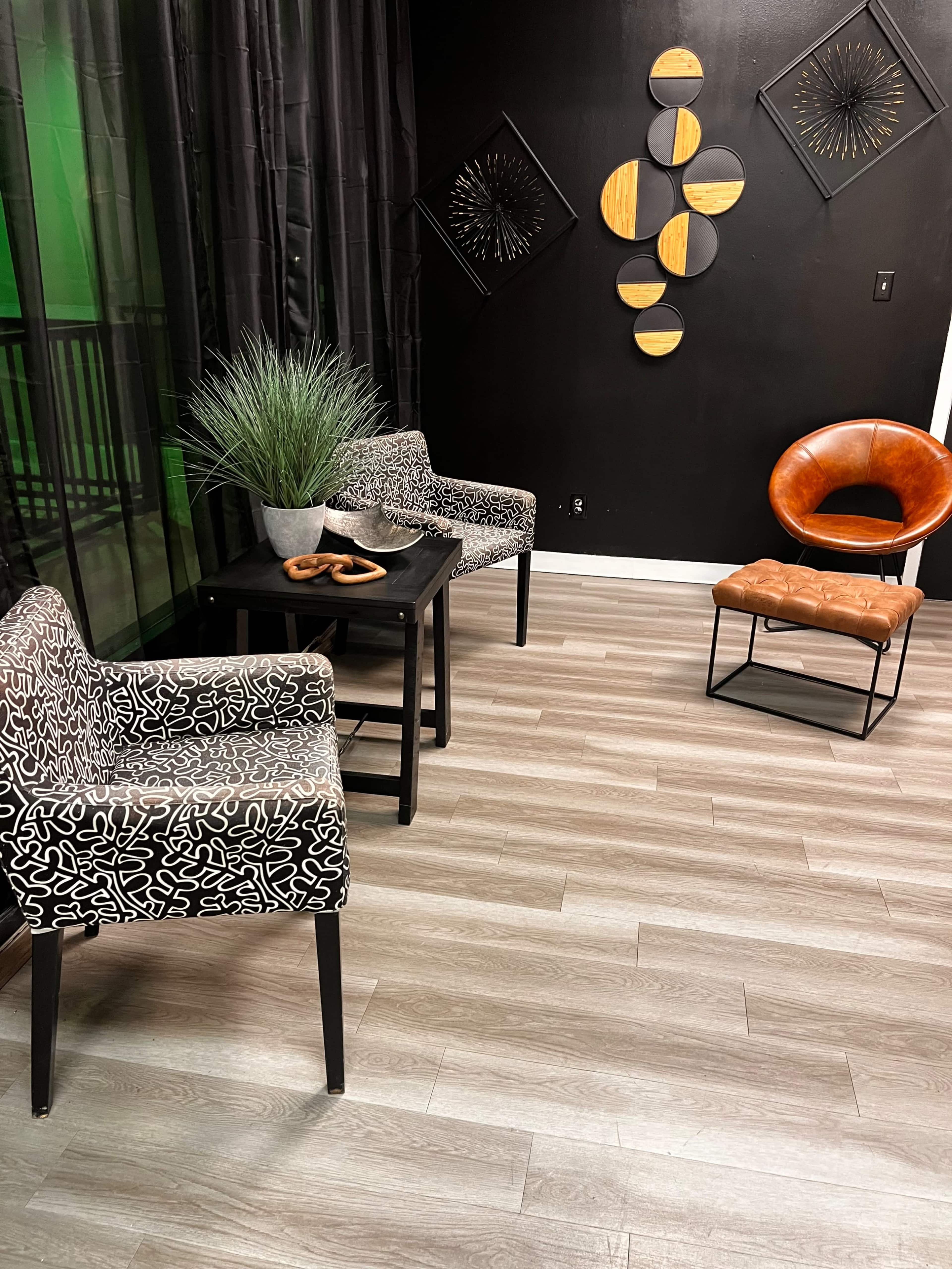 The image shows a cozy seating area with two patterned chairs, a small black side table, a brown chair, and decorative wall art against a dark backdrop.