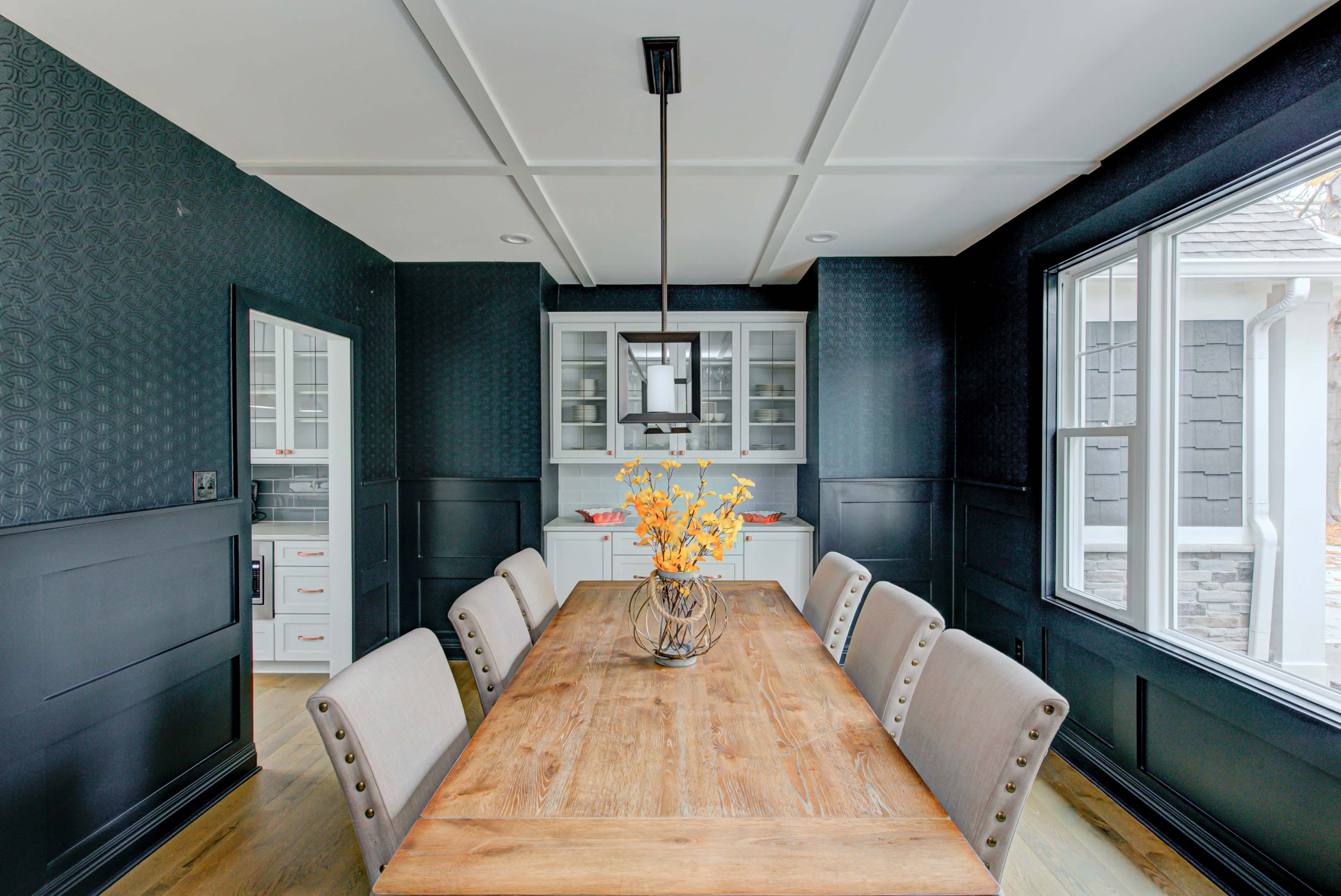 A dining room features a wooden table surrounded by upholstered chairs, with a modern light fixture and wall cabinetry in a dark blue setting.