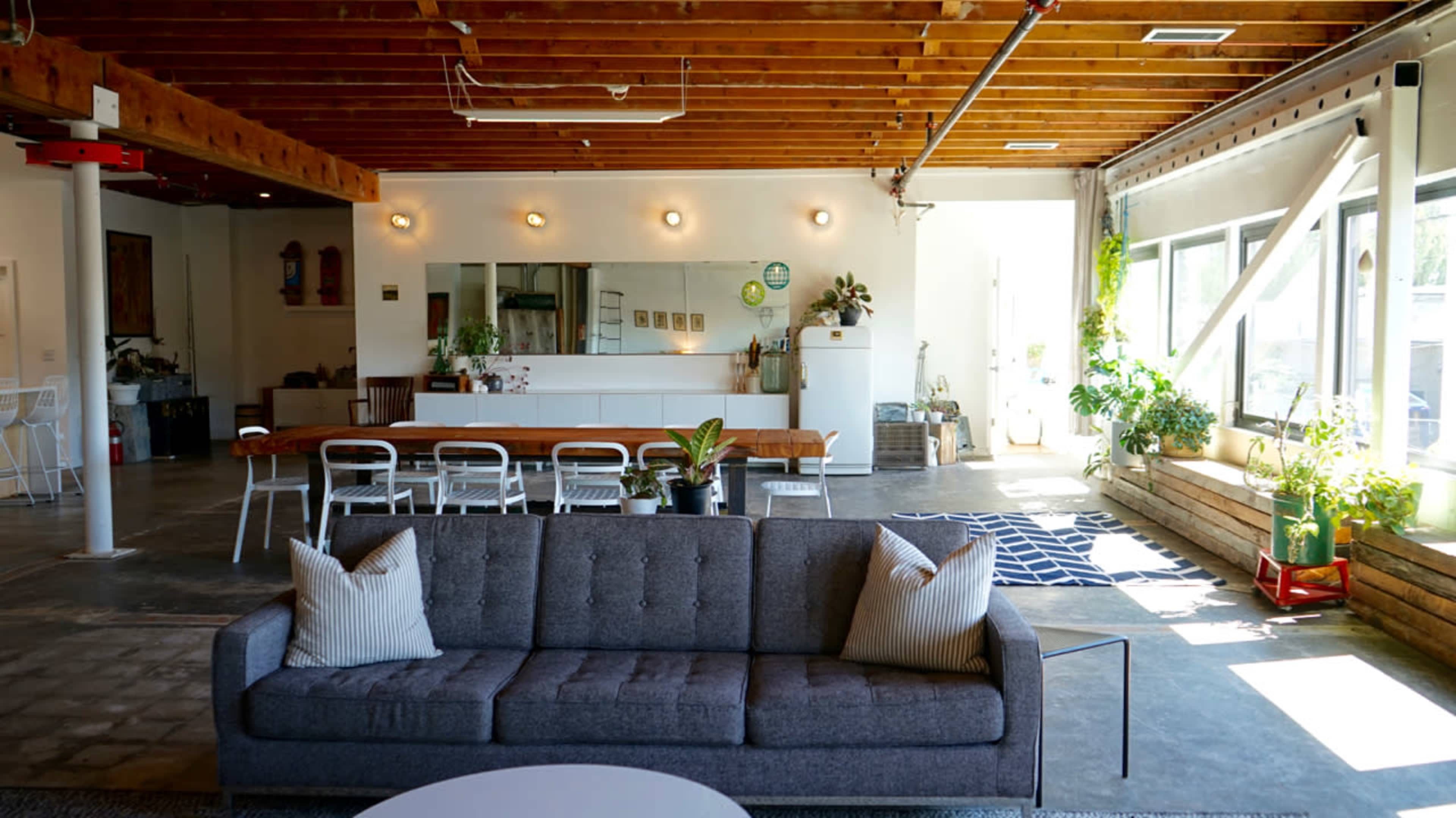 A spacious, modern interior with a gray sofa in the foreground, a long dining table in the center, and large windows allowing natural light to enter.