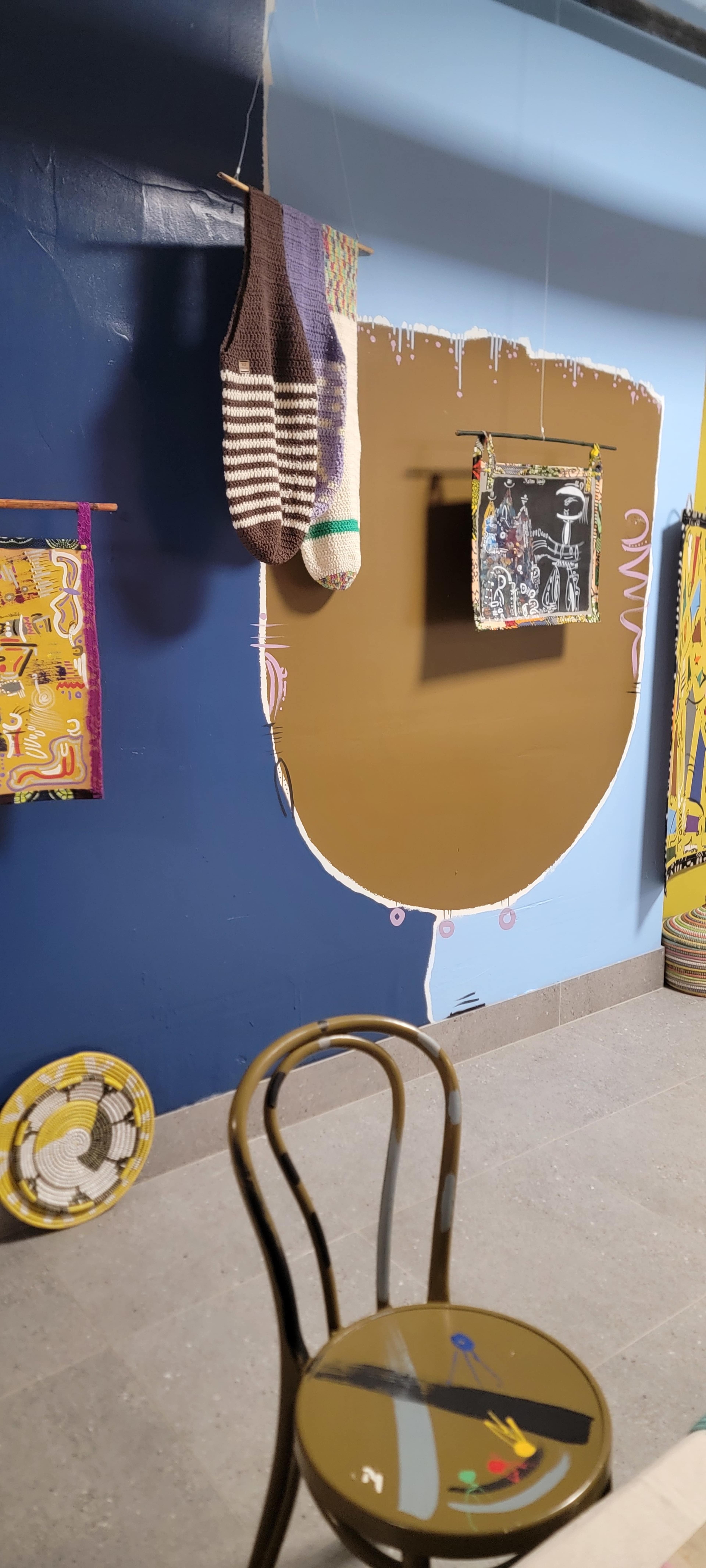 The image shows a room with a blue wall featuring a large brown oval shape painted on it, surrounded by various colorful artwork and a chair with a painted design.