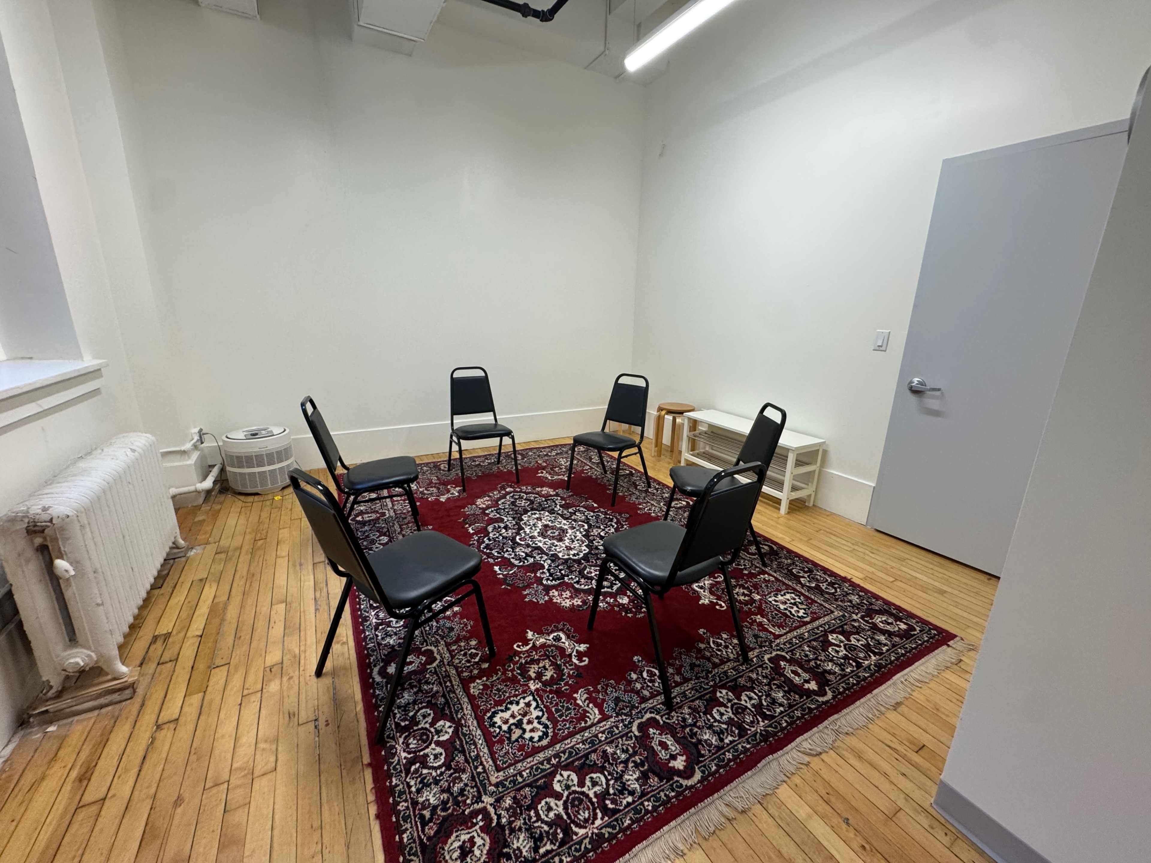 The image shows a small room with wooden floors, featuring a red patterned rug and six black chairs arranged in a circle.