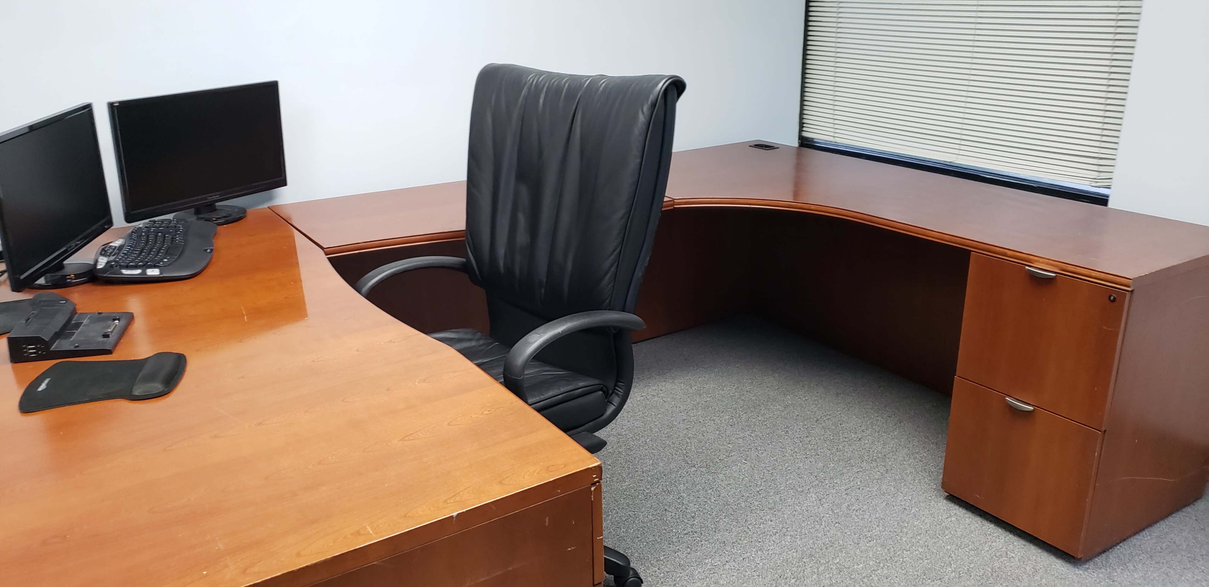 A simple office space featuring a wooden desk, a black leather office chair, two computer monitors, and a filing cabinet.