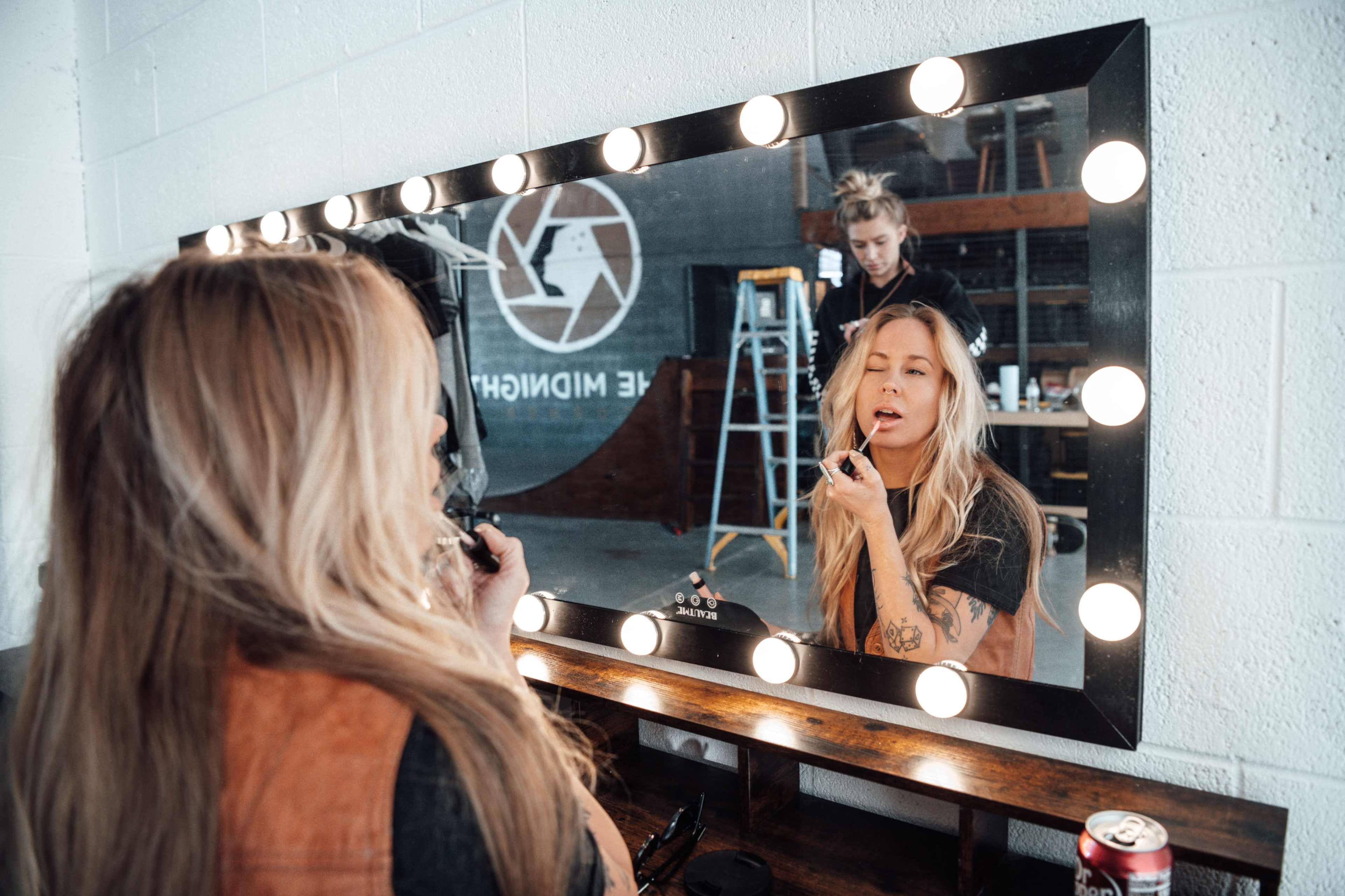 A woman applies lipstick while looking into a brightly lit mirror, with a second woman and a ladder visible in the background.