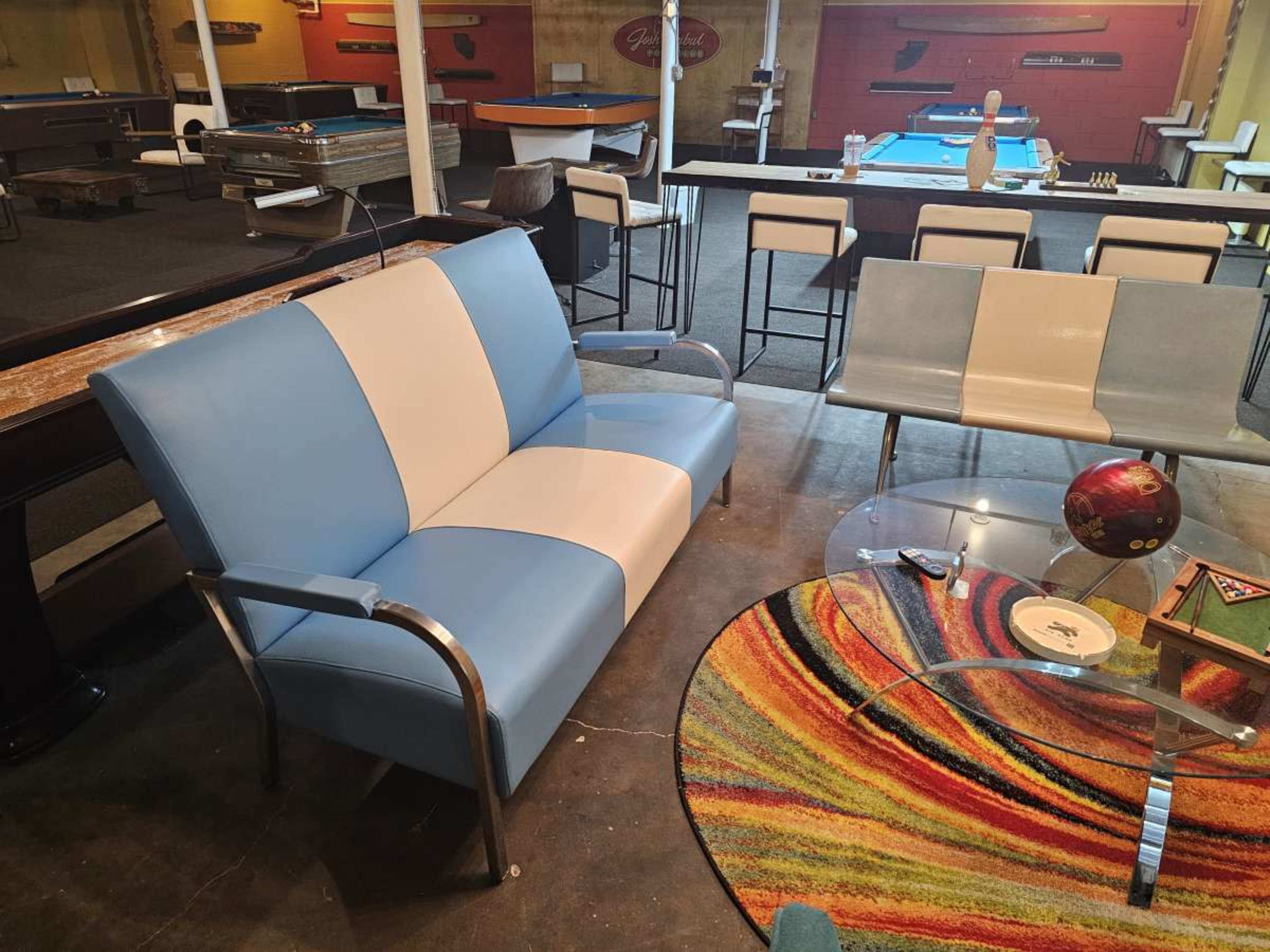 The image shows a modern blue and white two-tone loveseat in a game room filled with pool tables and seating areas.