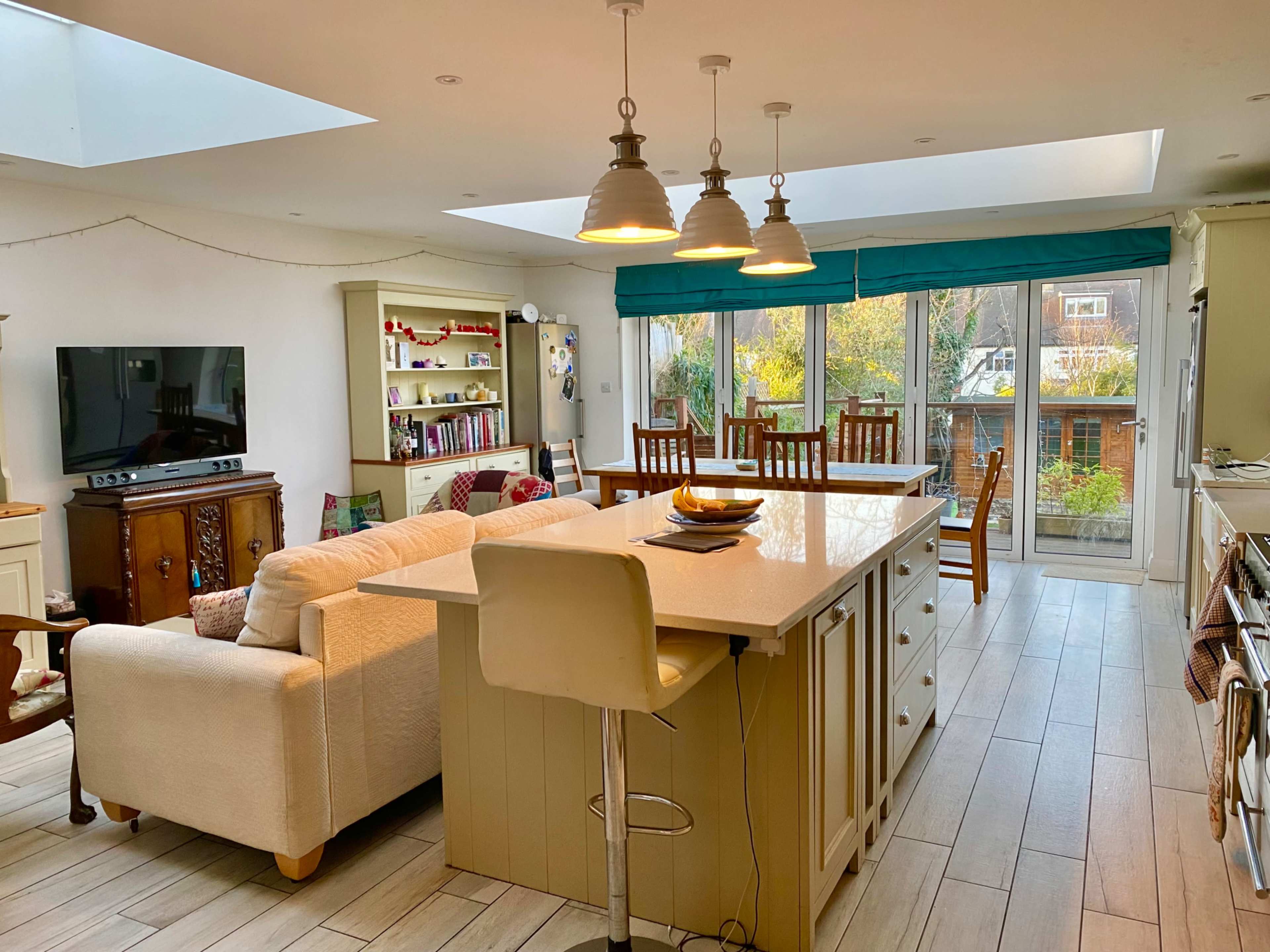 The image shows a bright, open-plan kitchen and living area featuring a large center island, seating at the counter, and large windows overlooking a garden.