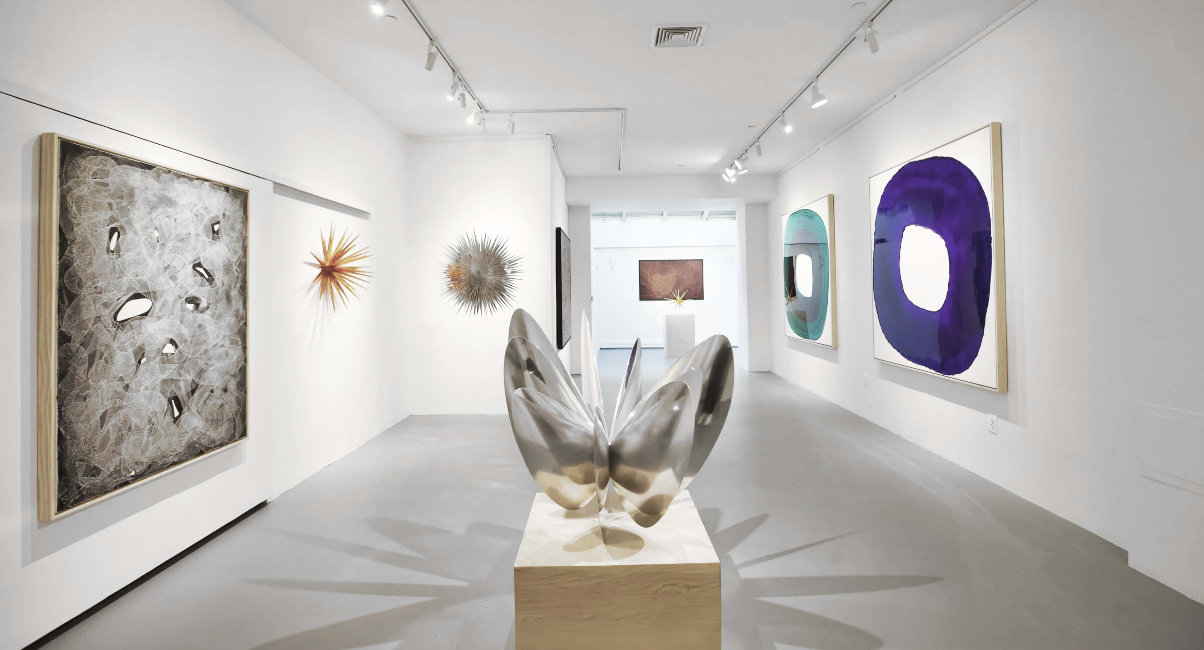 The image shows a contemporary art gallery with various abstract artworks displayed on the walls and a large metallic sculpture in the foreground.