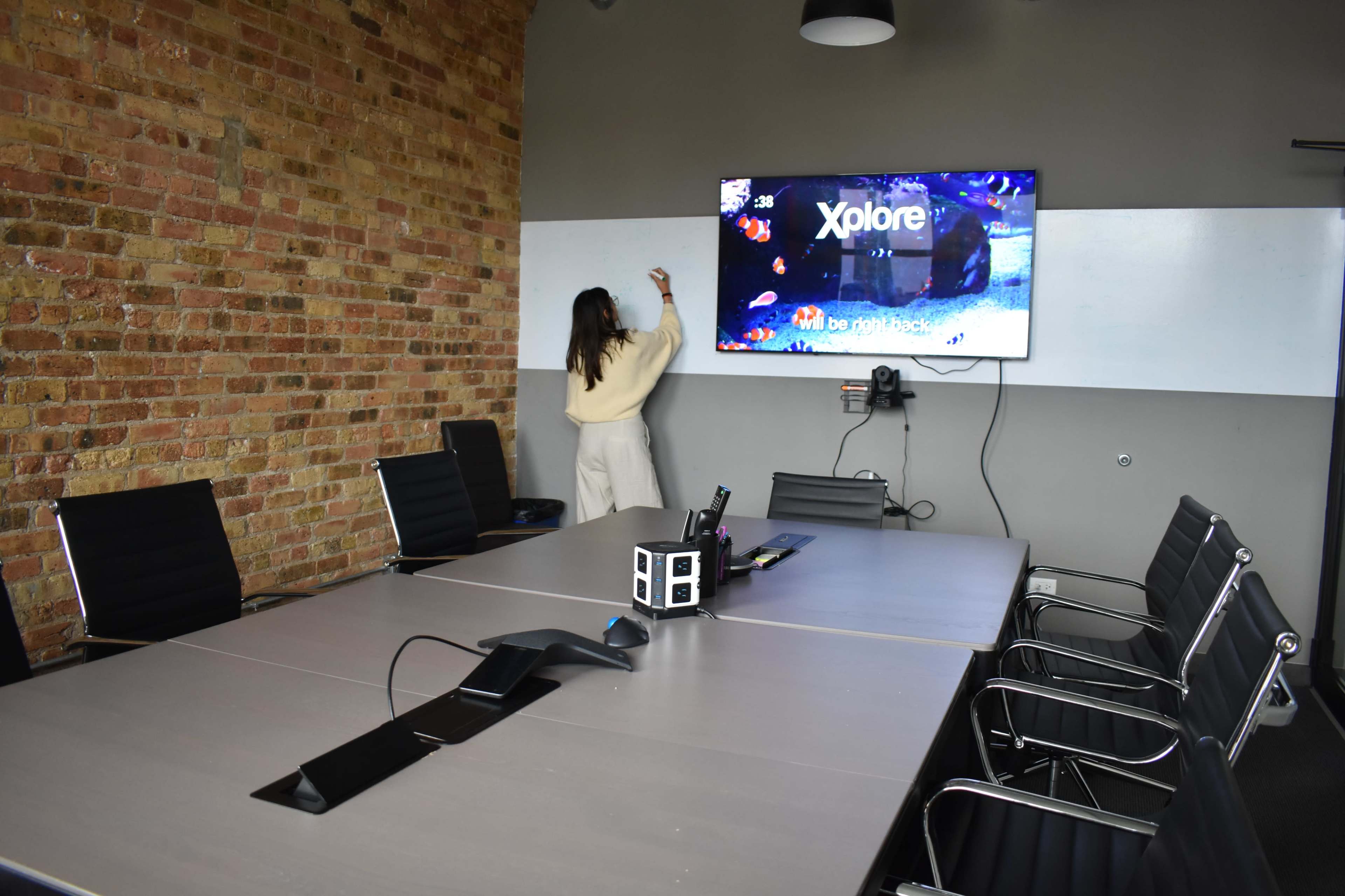 A woman stands in an empty conference room, writing on a screen displaying the word "Xplore," with a long table and black chairs arranged around it.