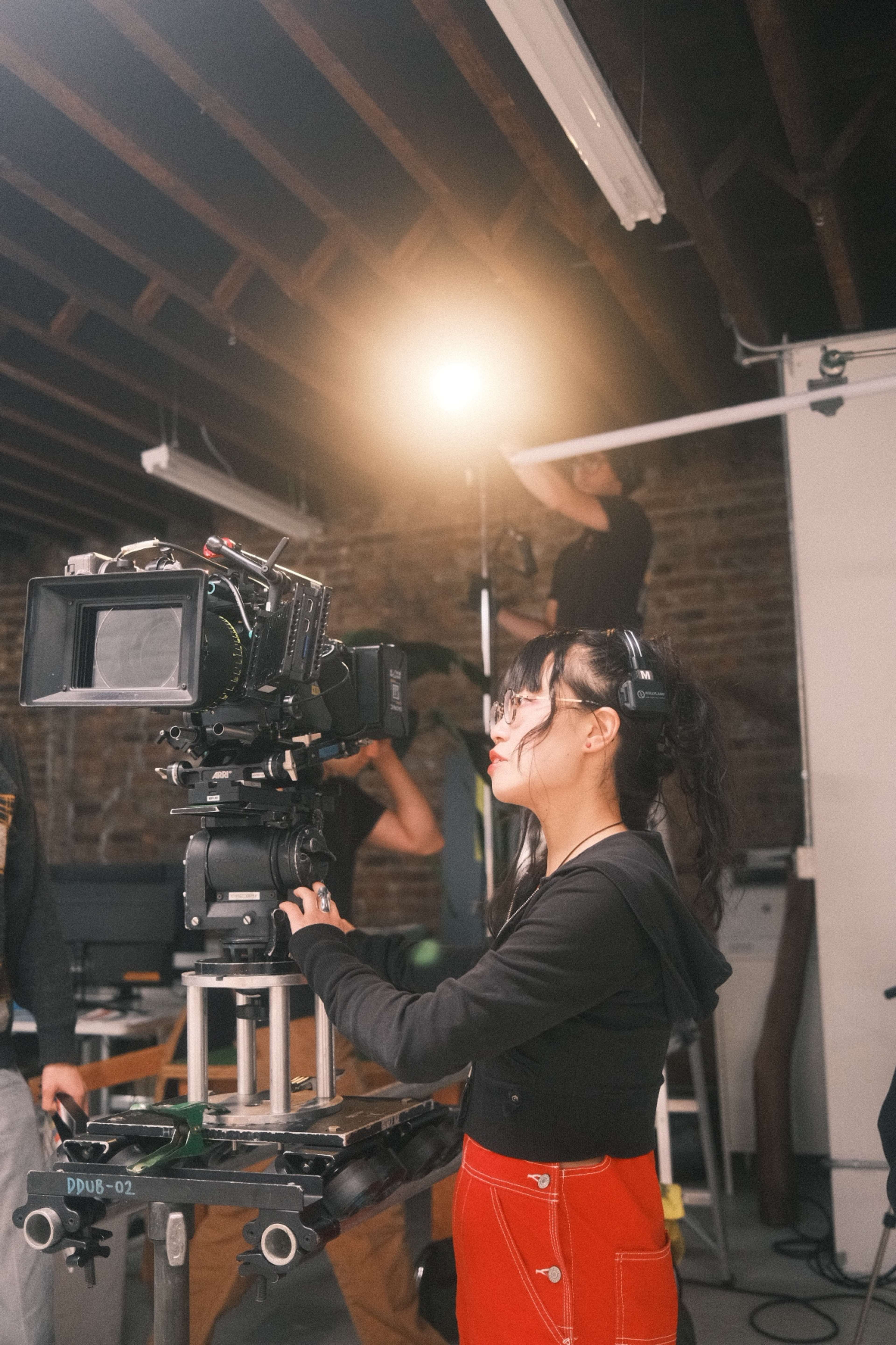 A woman operates a film camera on a tripod in an industrial-looking workspace while another person adjusts a light in the background.