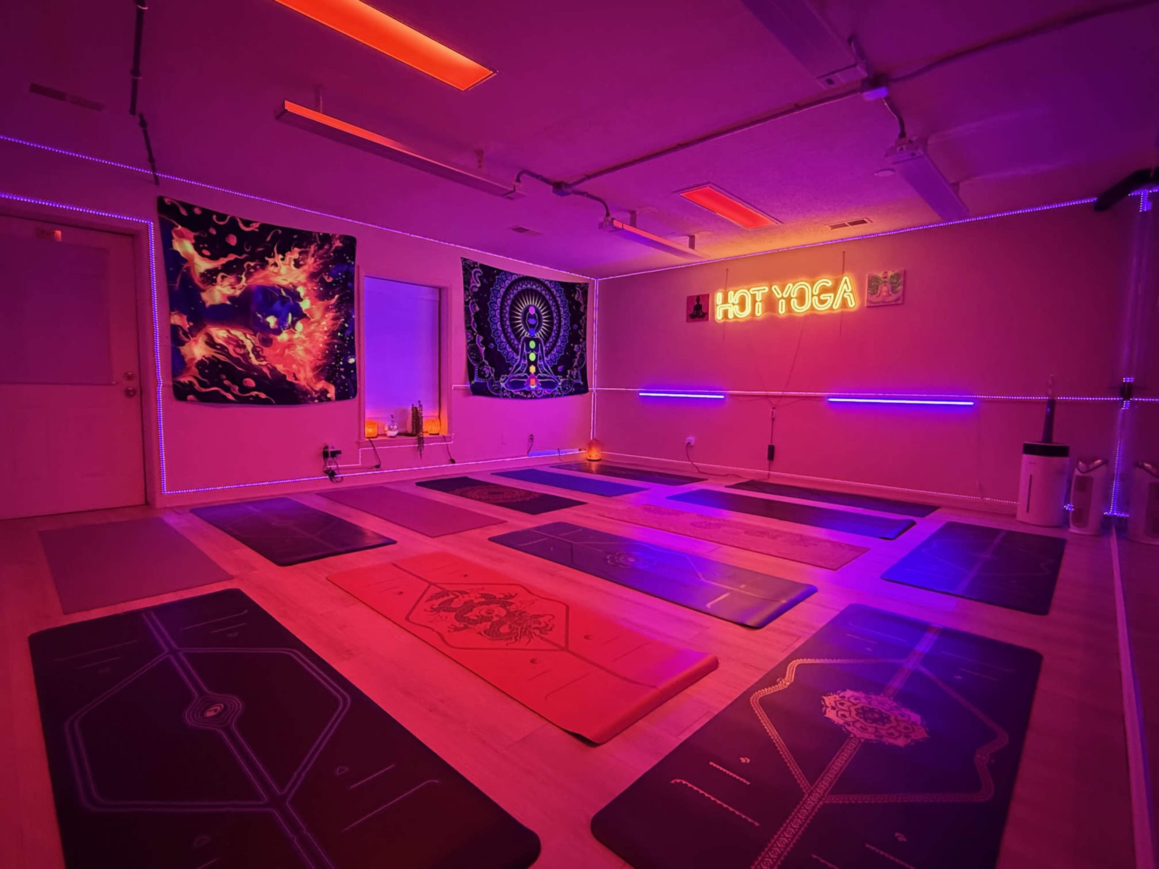 The image depicts a dimly lit hot yoga studio with several yoga mats arranged on the floor and colorful neon lighting illuminating the space.