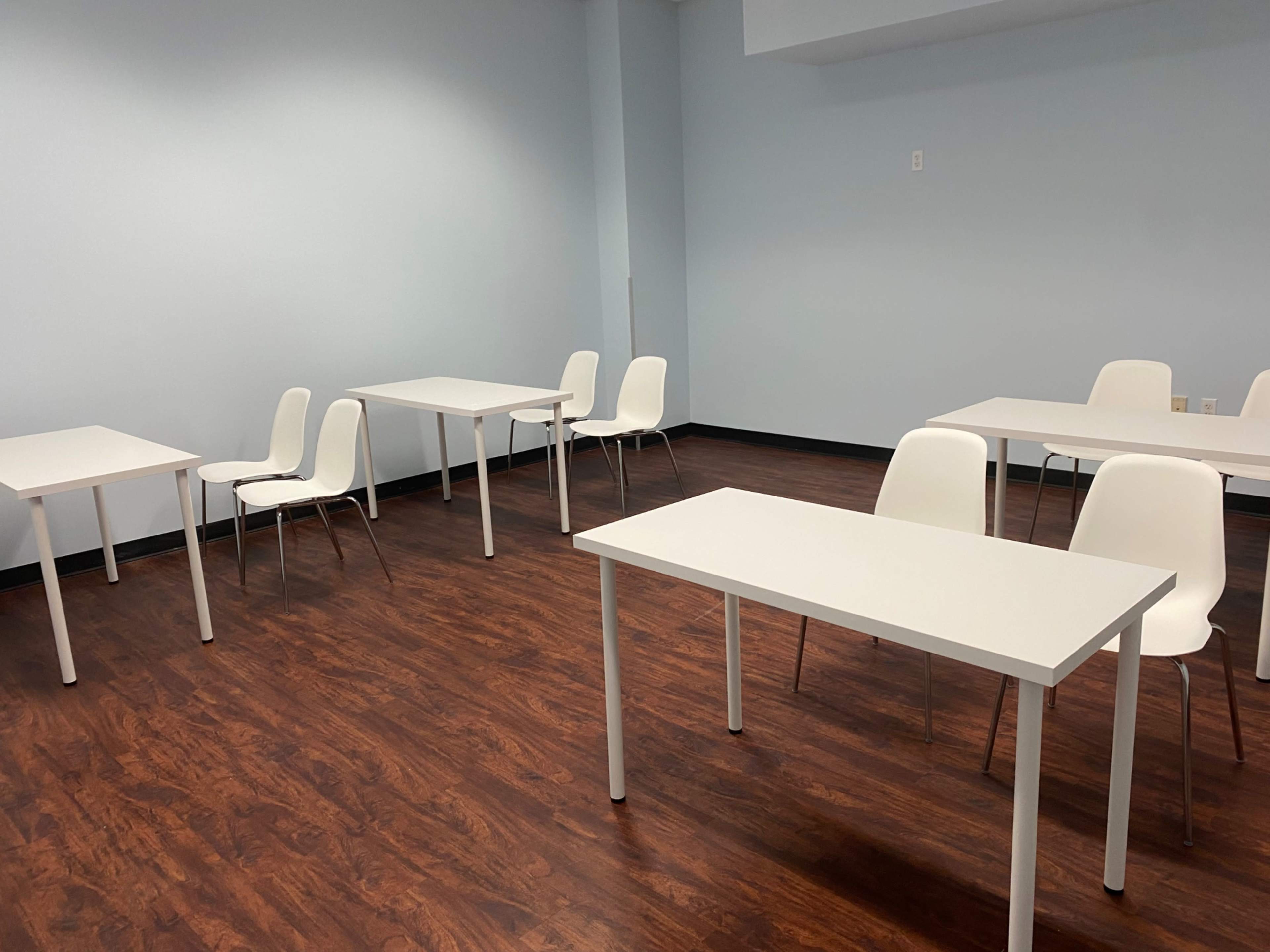 A minimalistic classroom features several white tables and chairs arranged in a spacious, well-lit area with wooden flooring.