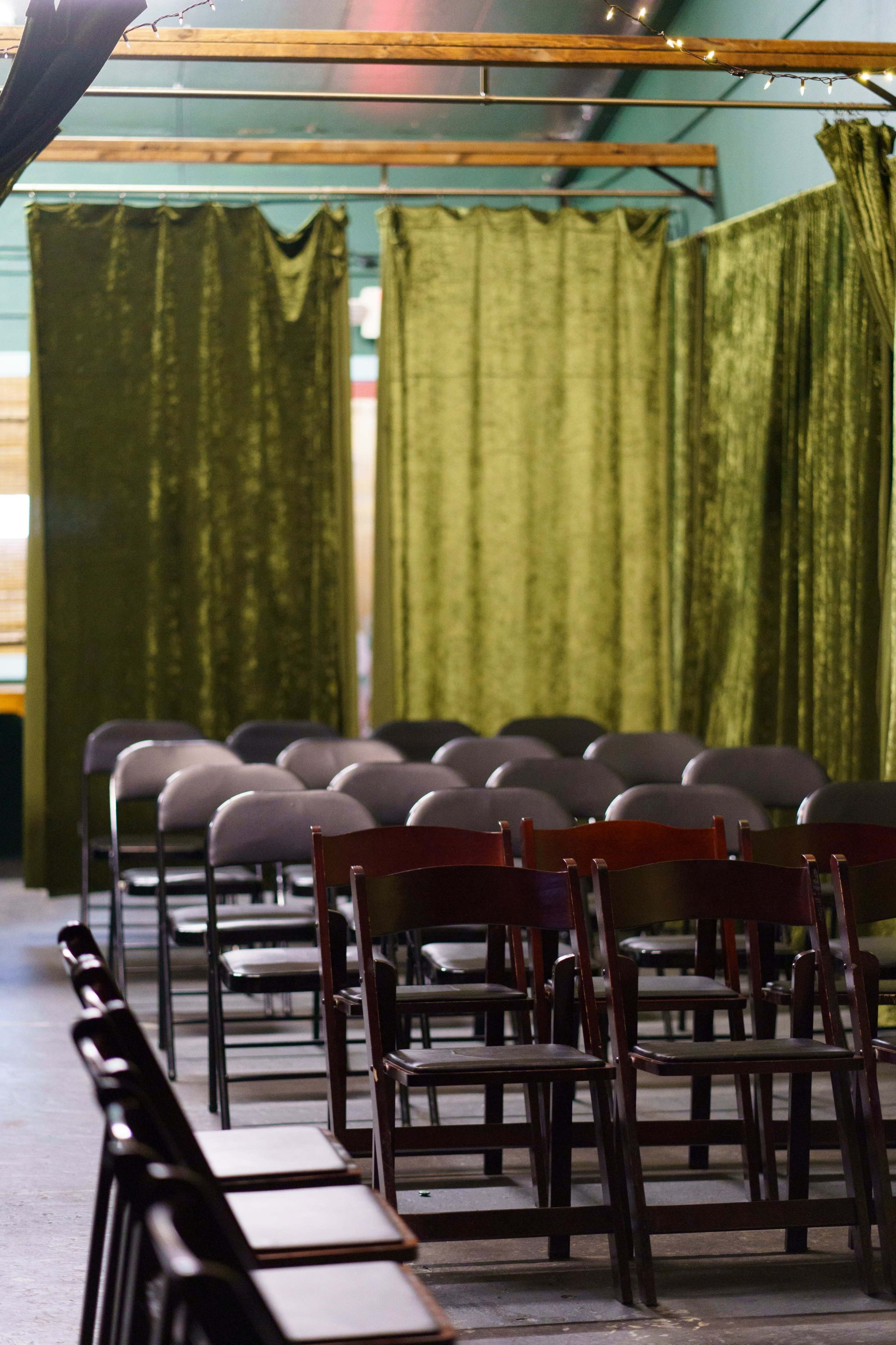 The image shows a seating arrangement with rows of gray chairs facing a green velvet curtain in a dimly lit indoor space.