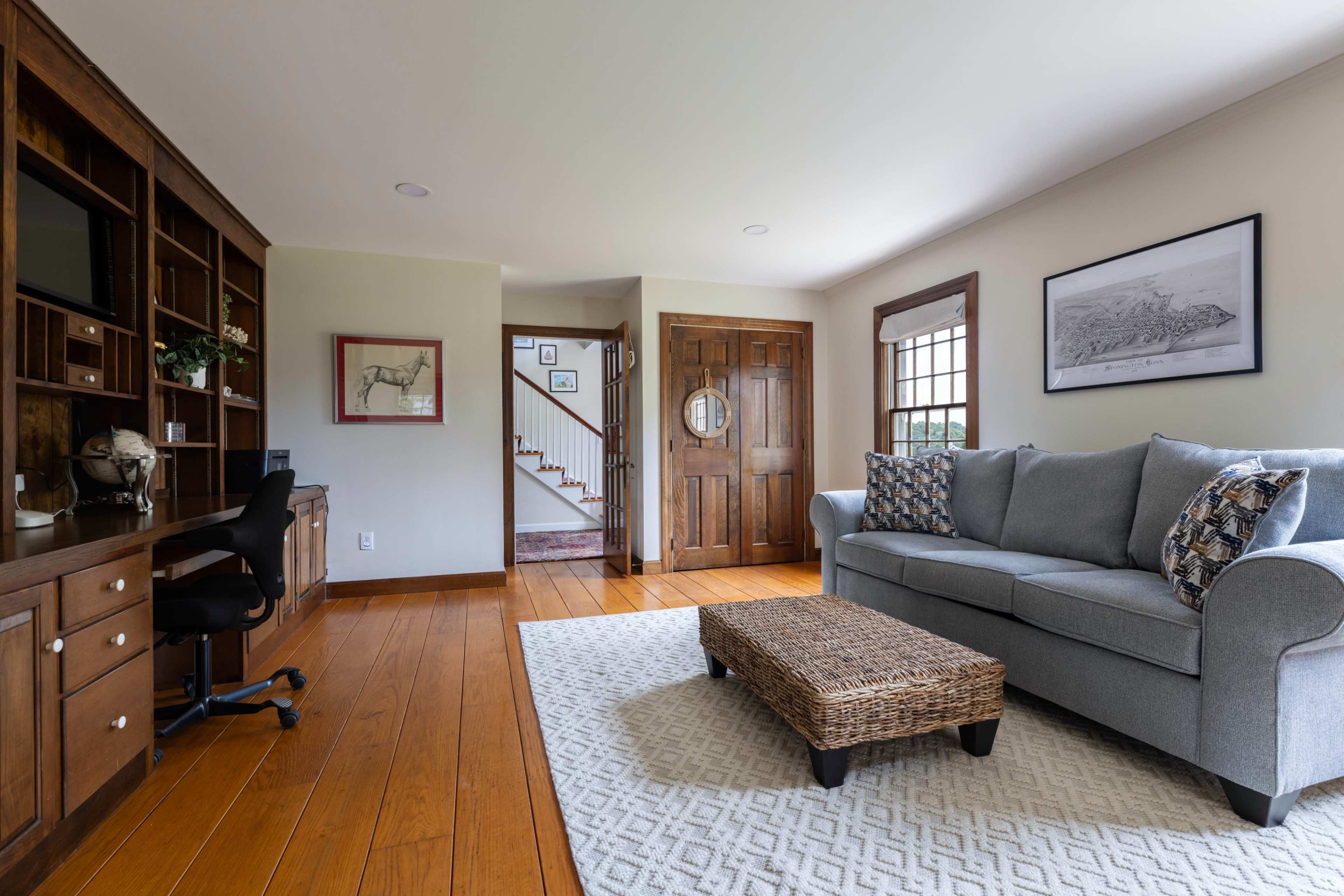 The image shows a living room with a gray couch, a woven coffee table, a wooden desk, and a staircase leading to another level.