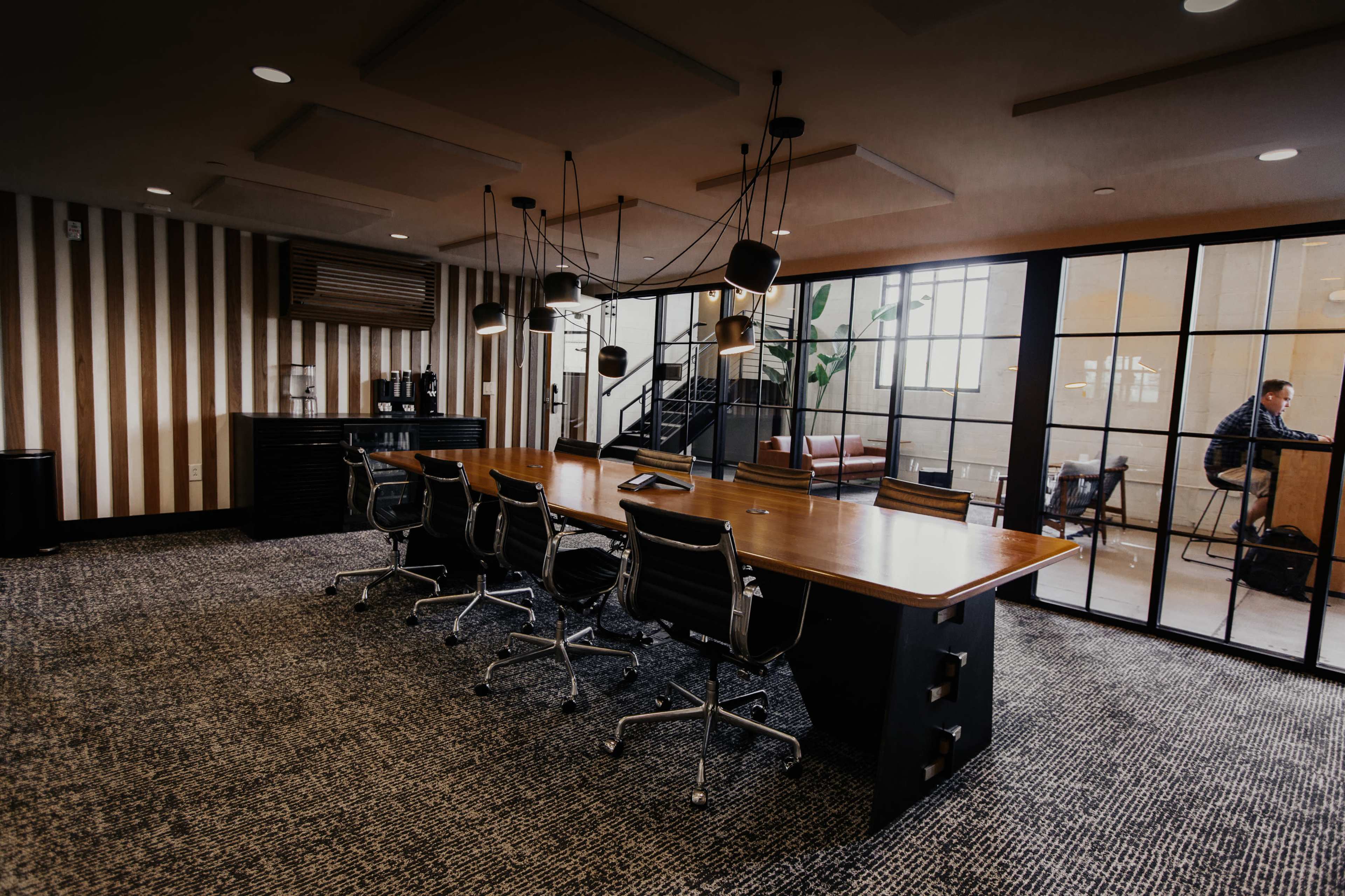 A modern meeting room features a large wooden table surrounded by several black office chairs, with a coffee station and large windows in the background.
