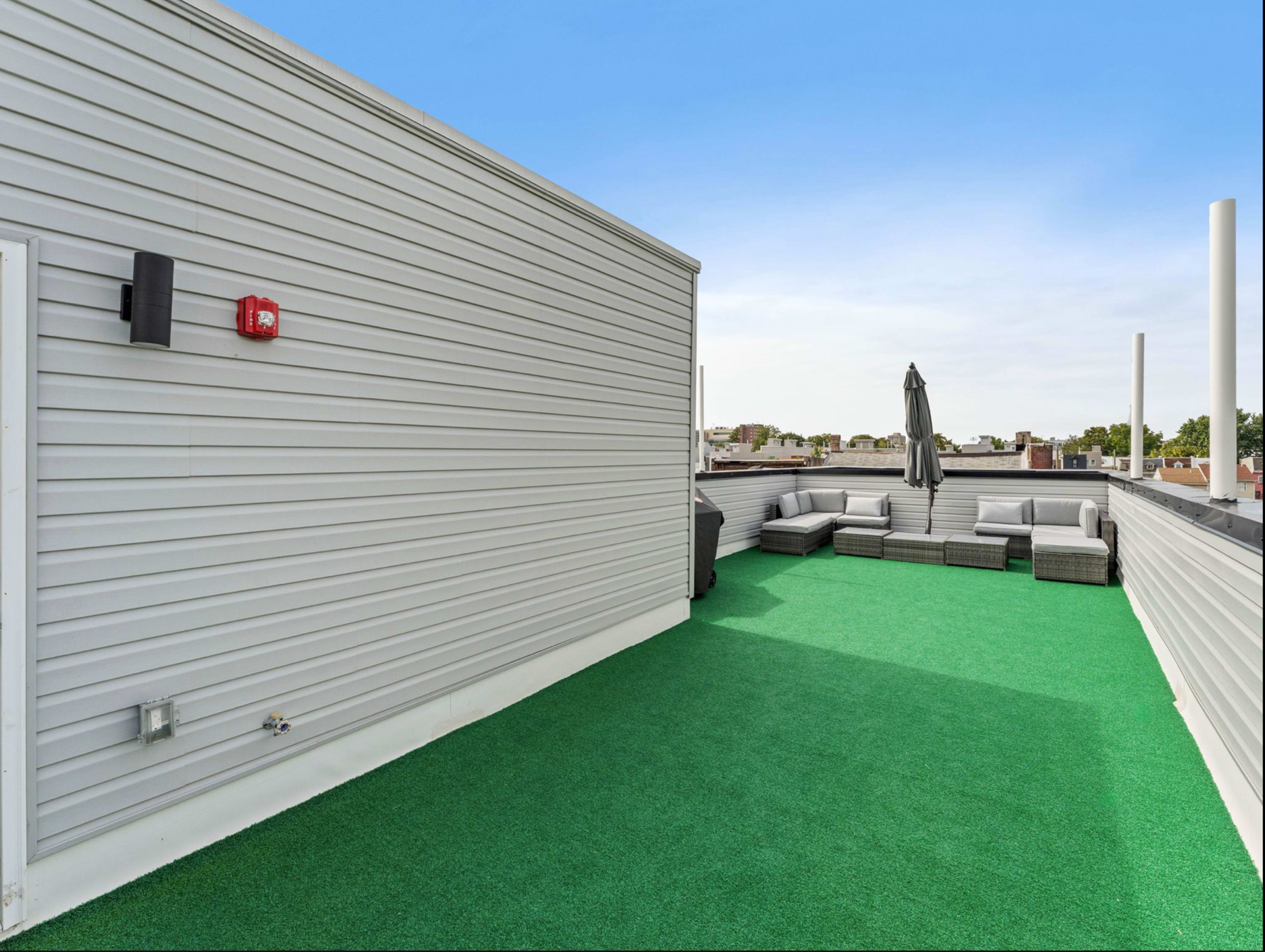 A rooftop space features artificial grass flooring, a lounge area with gray outdoor furniture, and an umbrella.