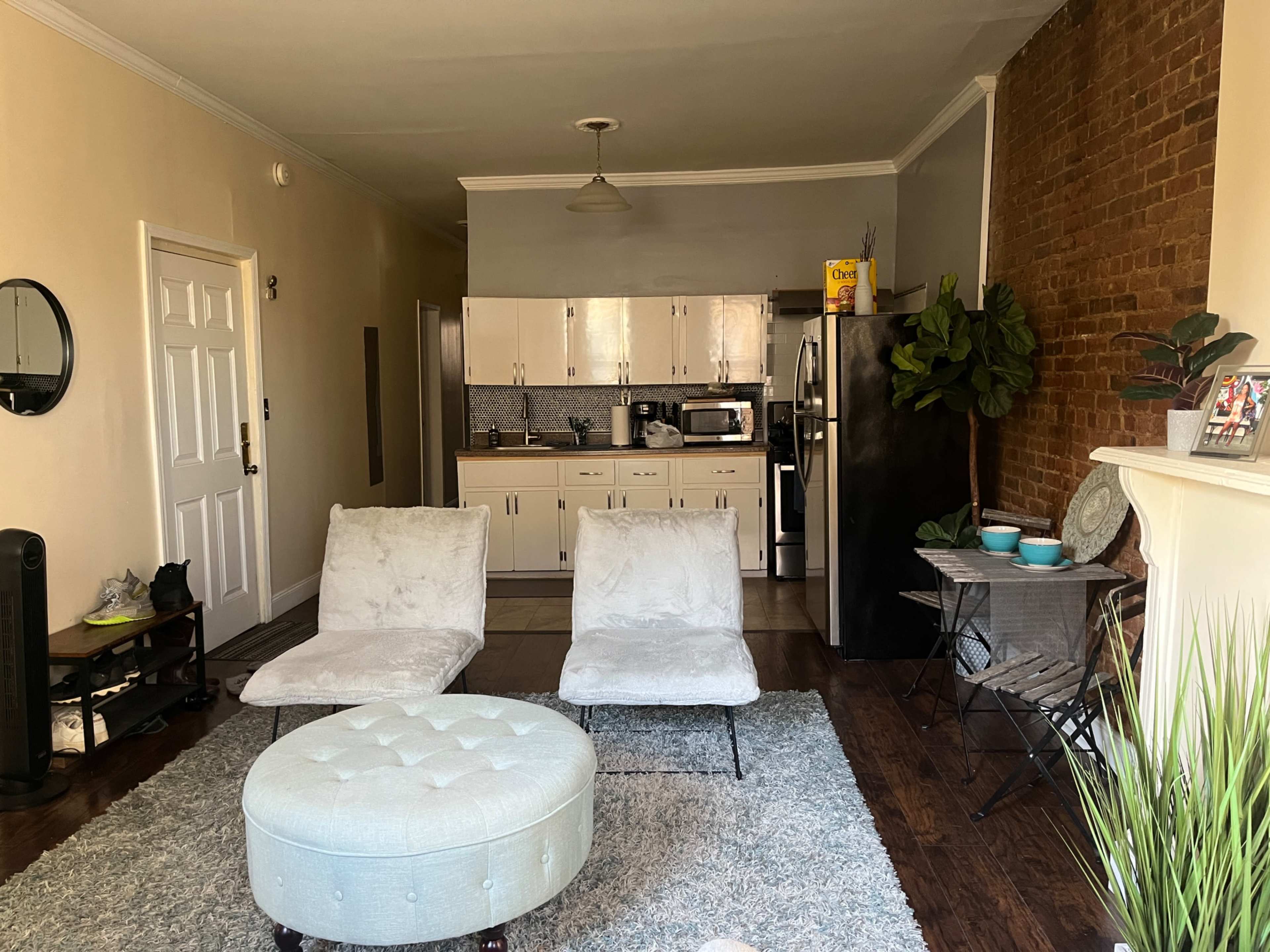A living room with two armchairs, an ottoman, a small dining table, kitchen area, and a brick wall.