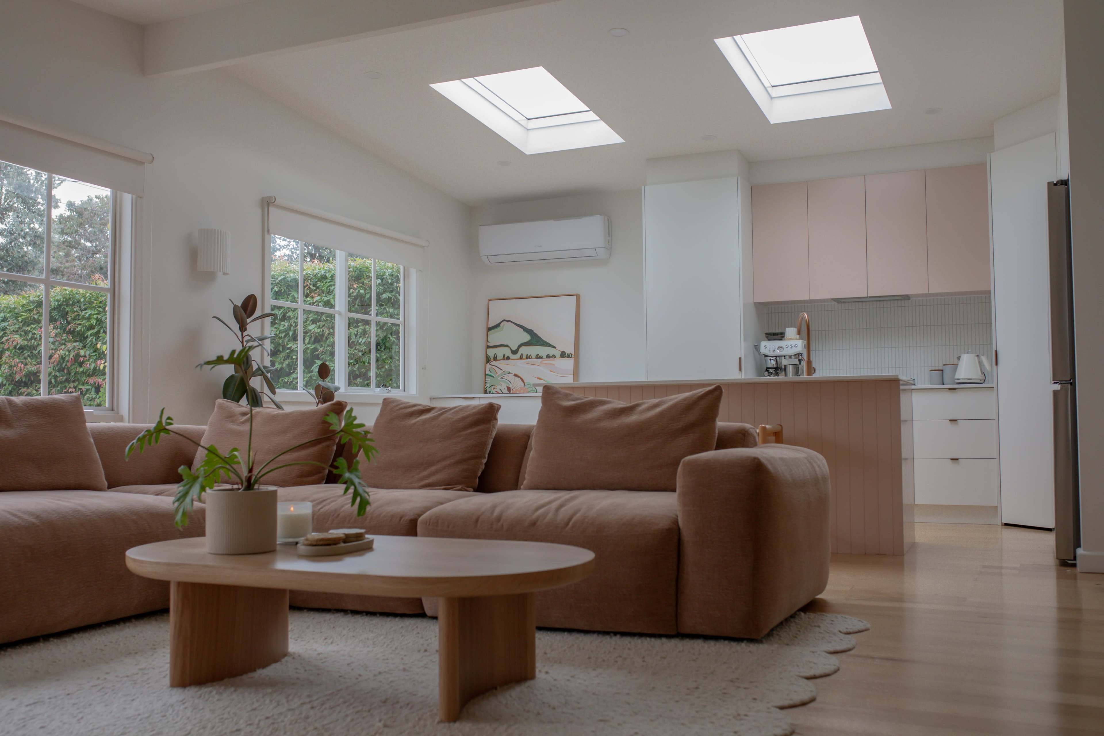 The image shows a modern, open living space with a large beige sofa, a circular wooden coffee table, and skylights illuminating the room.