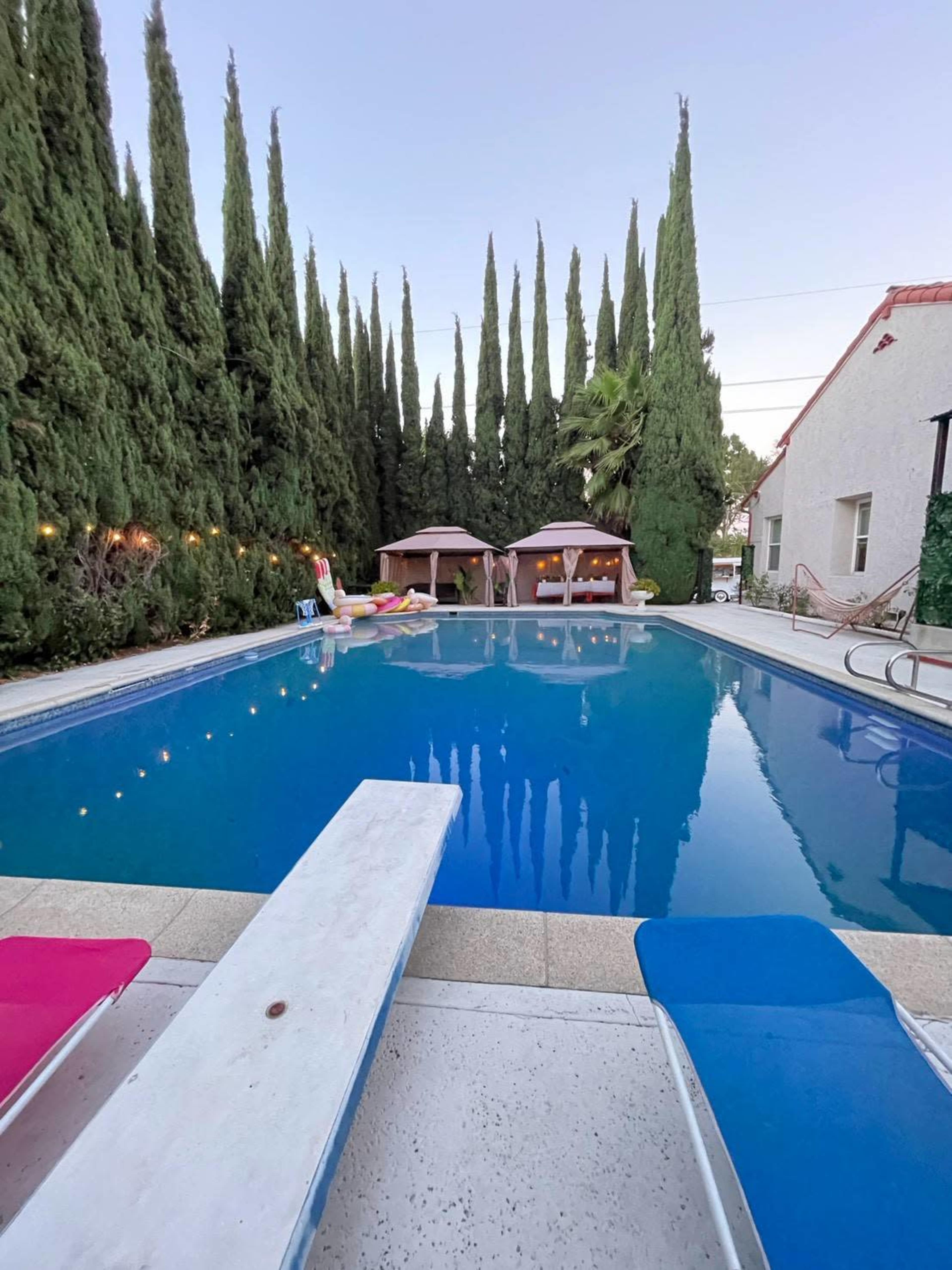 The image shows a swimming pool surrounded by tall conifer trees and two pink cabanas.