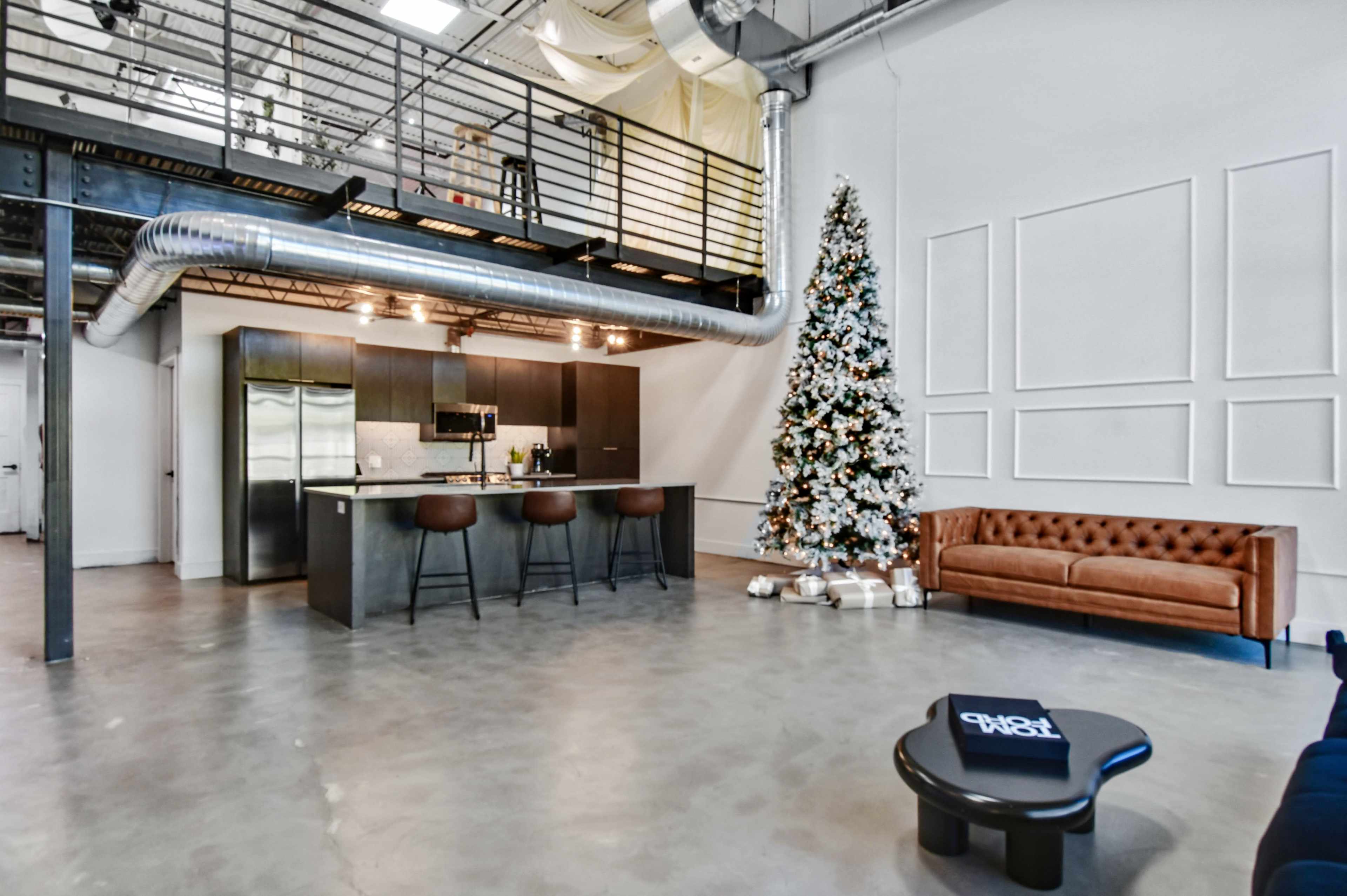 The image shows a modern, industrial-style loft featuring a cozy living area with a brown leather sofa, a minimalist coffee table, a kitchen with stainless steel appliances, and a decorated Christmas tree in the corner.