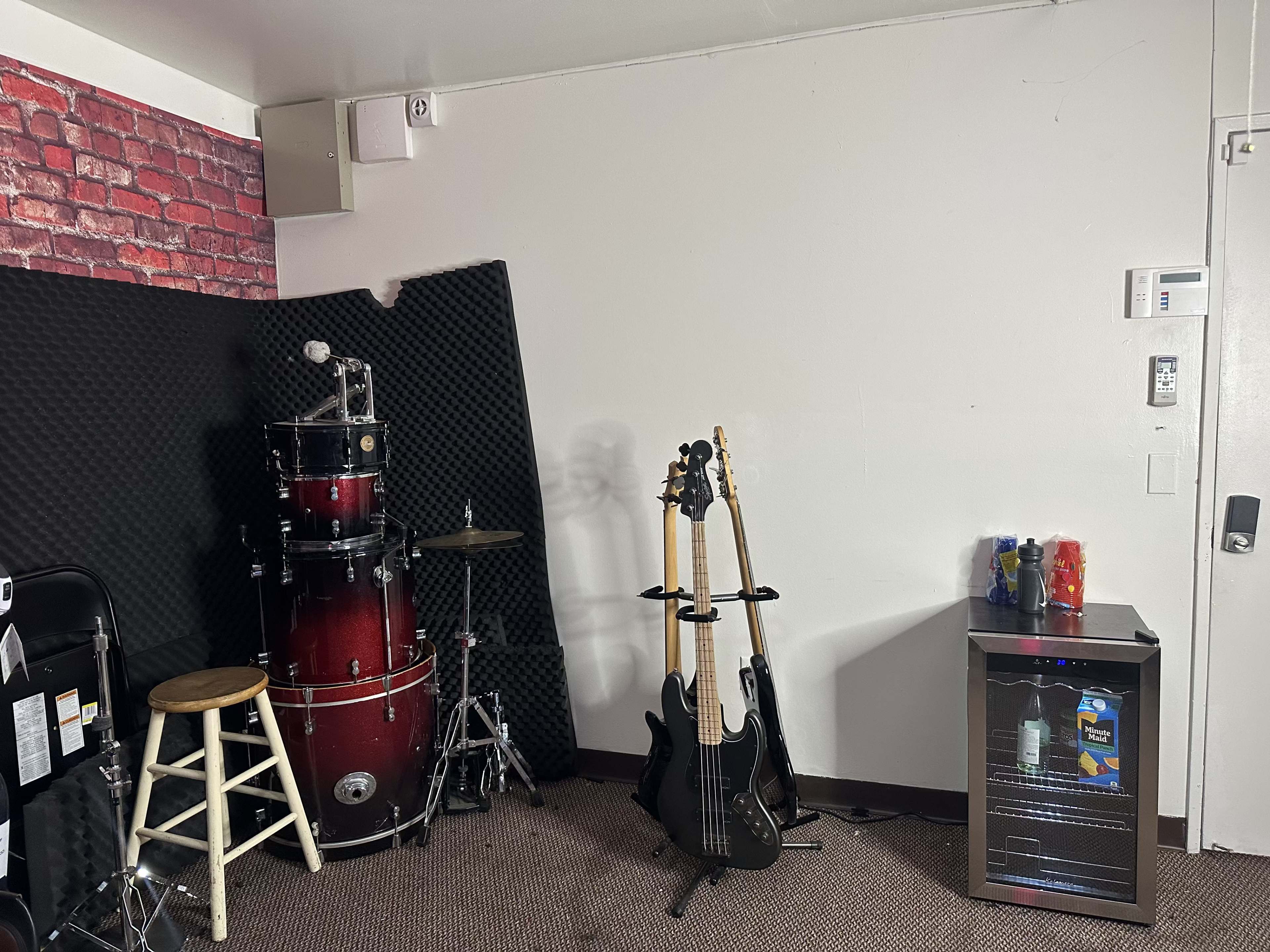 The image shows a practice space with a drum set, two electric guitars on stands, and a mini fridge against a textured wall.