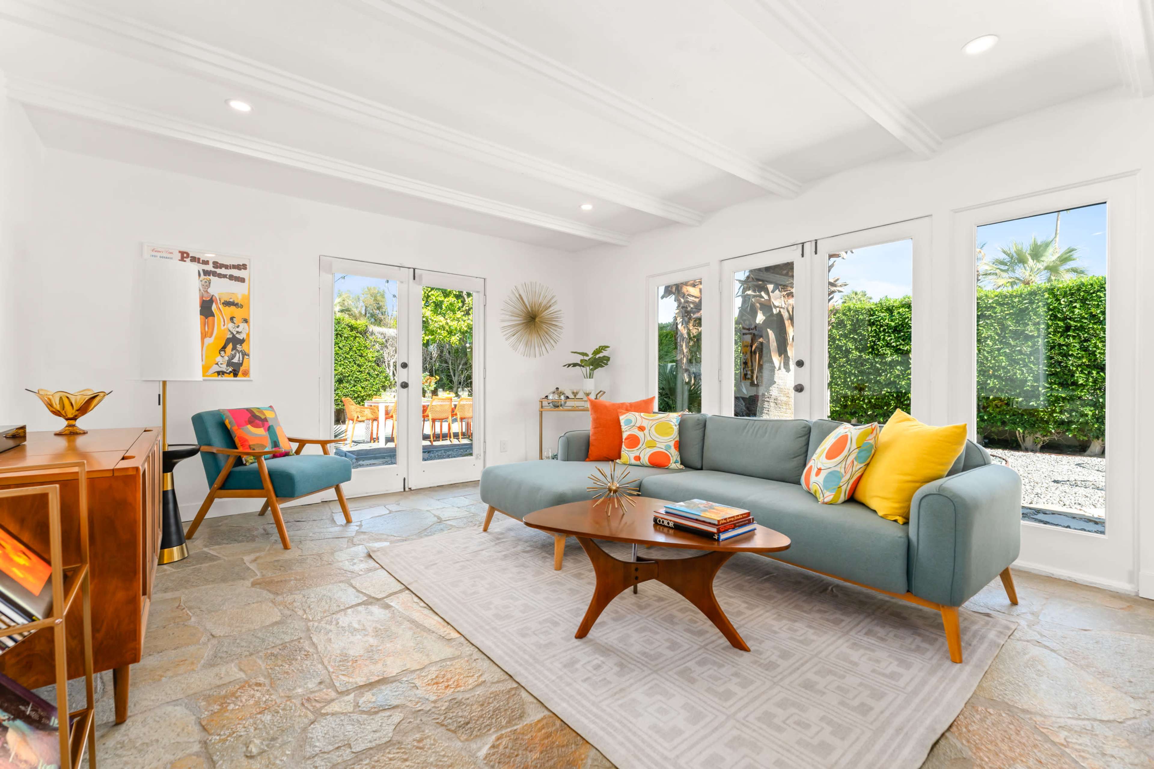 A bright living room features a blue couch with colorful pillows, a wooden coffee table, and large windows opening to a landscaped outdoor space.