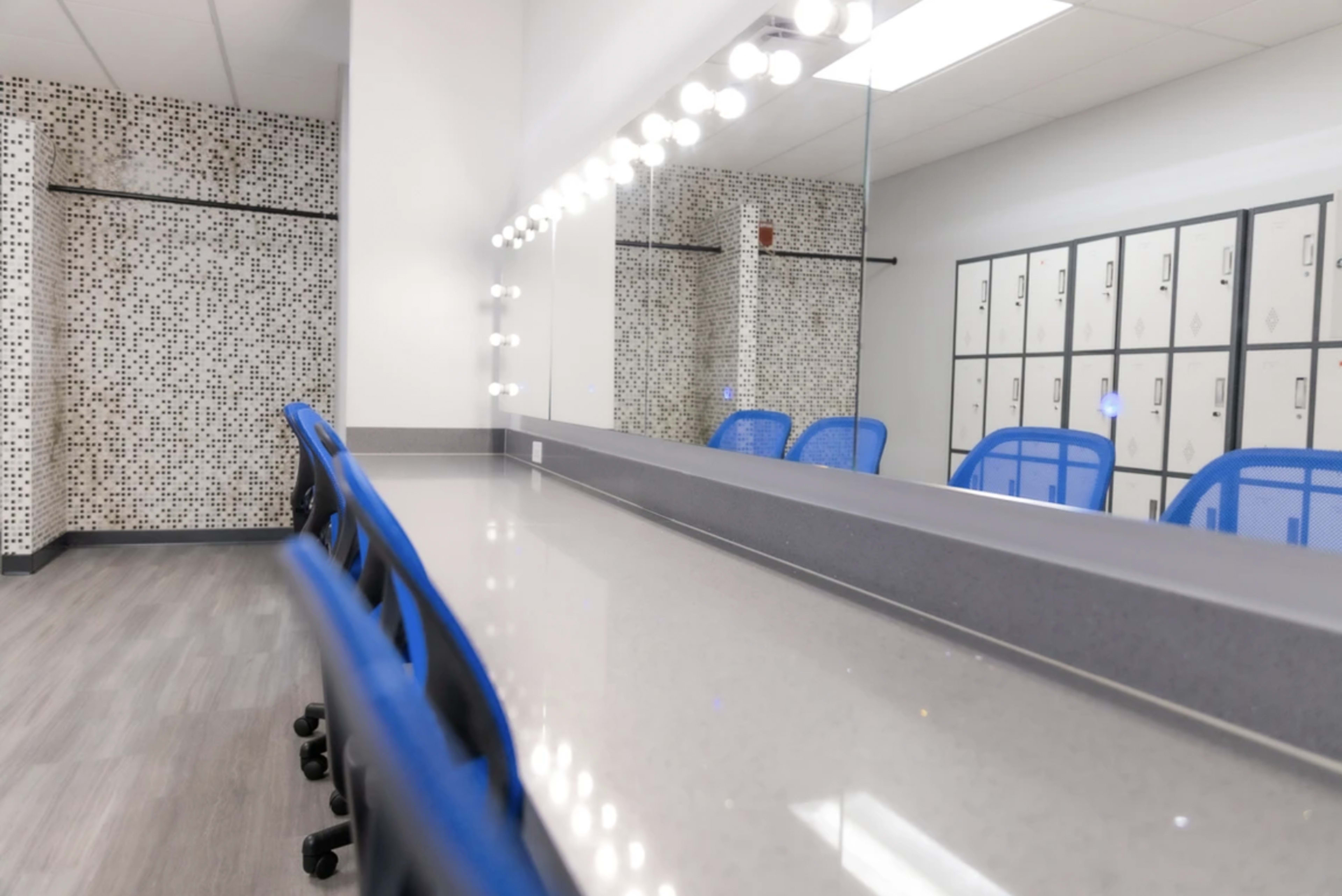A mirrored vanity area with bright lights and blue chairs beside a row of lockers against a patterned wall.