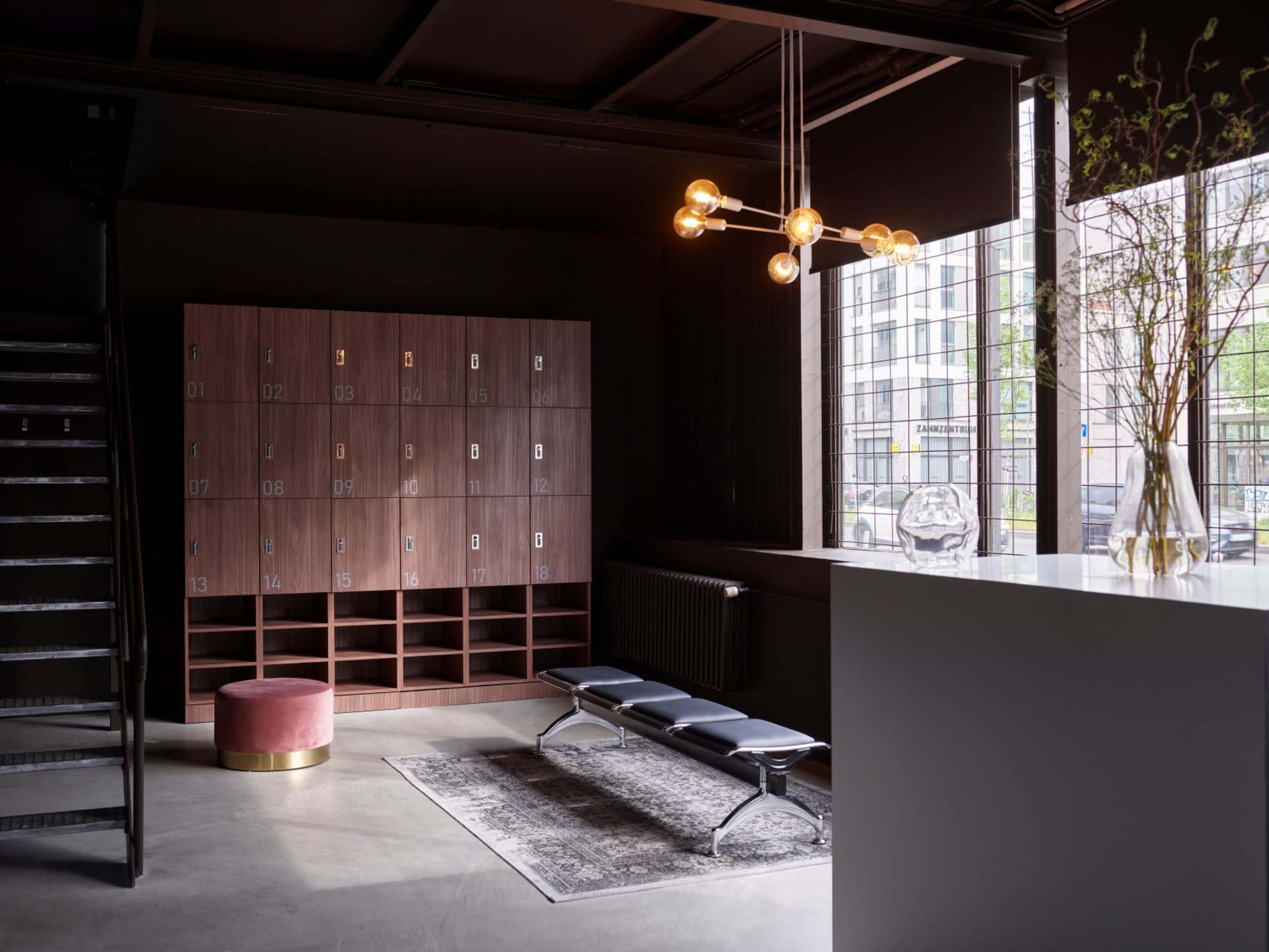 A stylish lobby area featuring a wooden locker unit, a round upholstered ottoman, a sleek bench, and a large window with natural light illuminating the space.