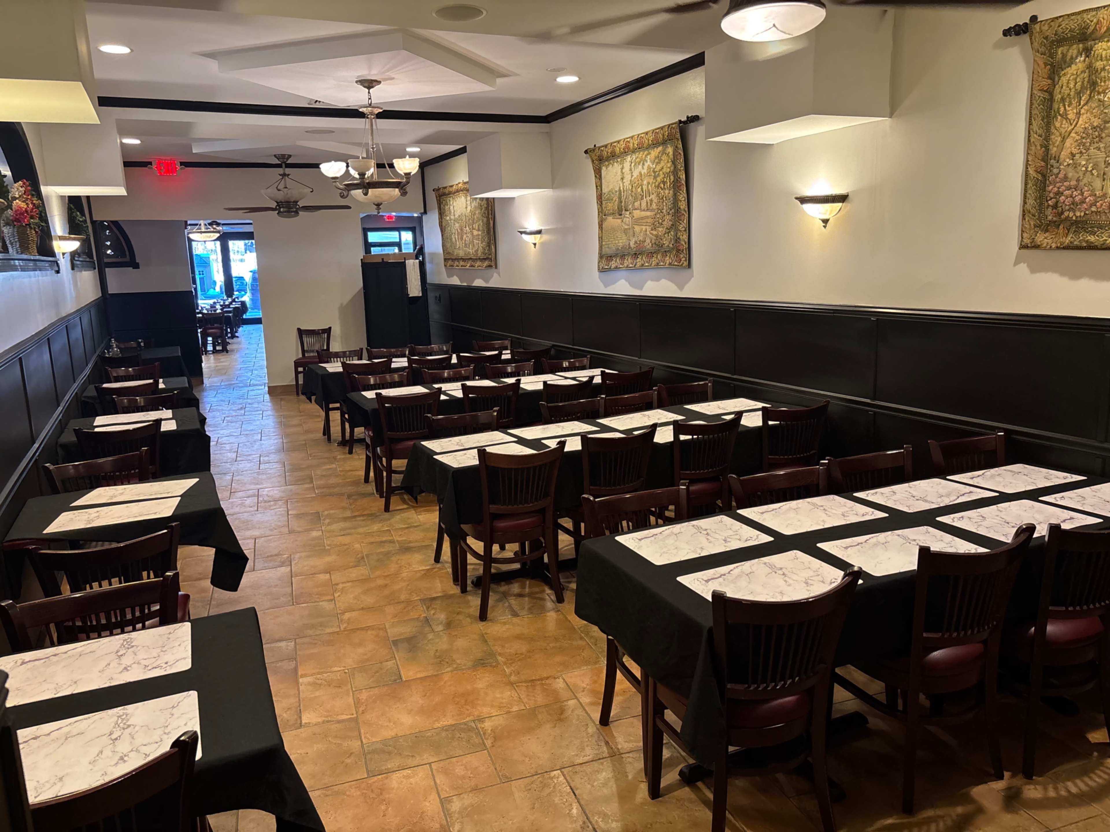 The image shows a long, narrow restaurant interior with multiple tables covered in black tablecloths, set with marble tops, and complemented by decorative wall hangings.