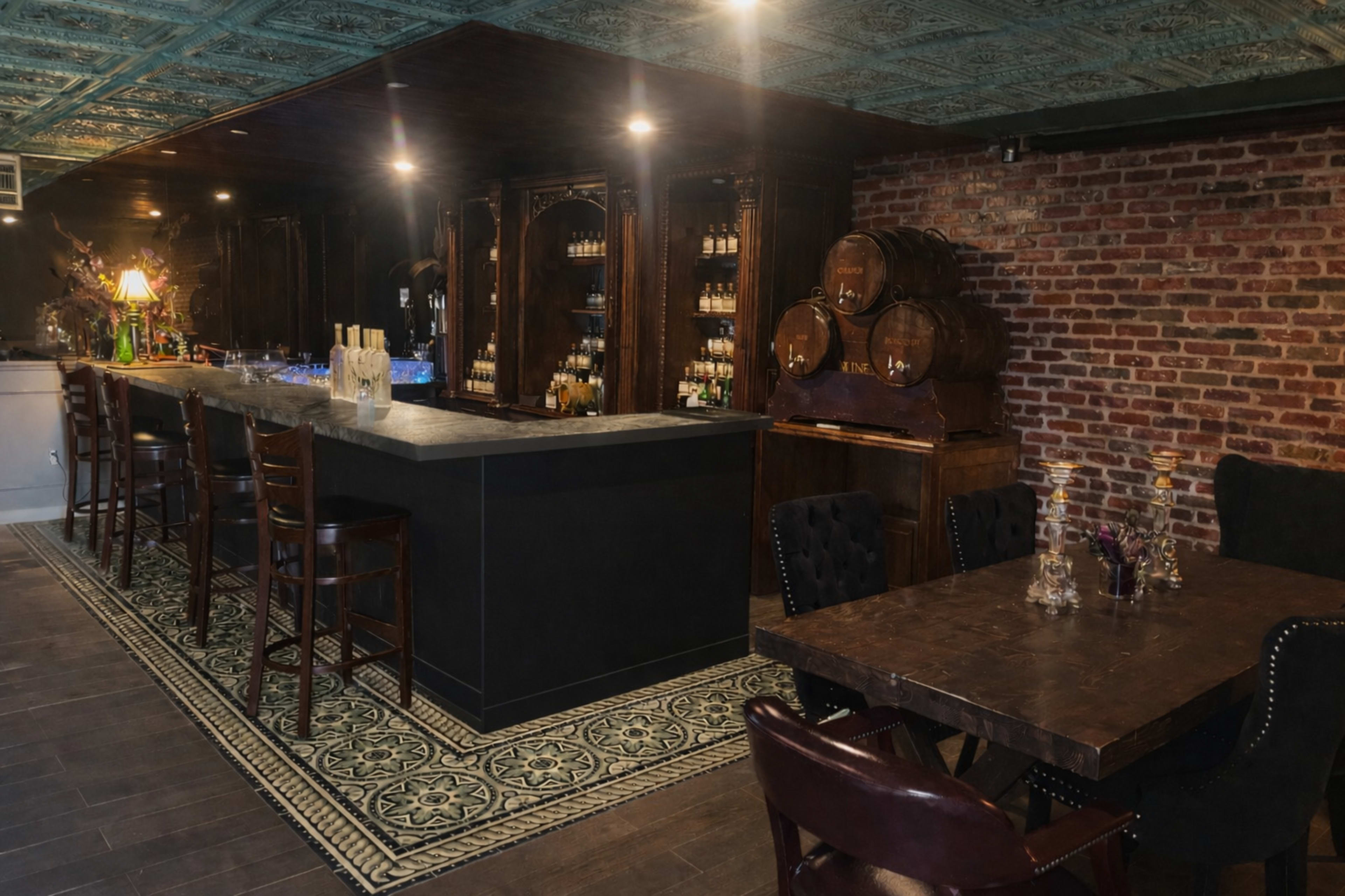The image shows an elegant bar area with a dark wood counter, shelves stocked with various bottles, and a brick wall backdrop, accompanied by wooden barrels and a seating area with dark furniture.