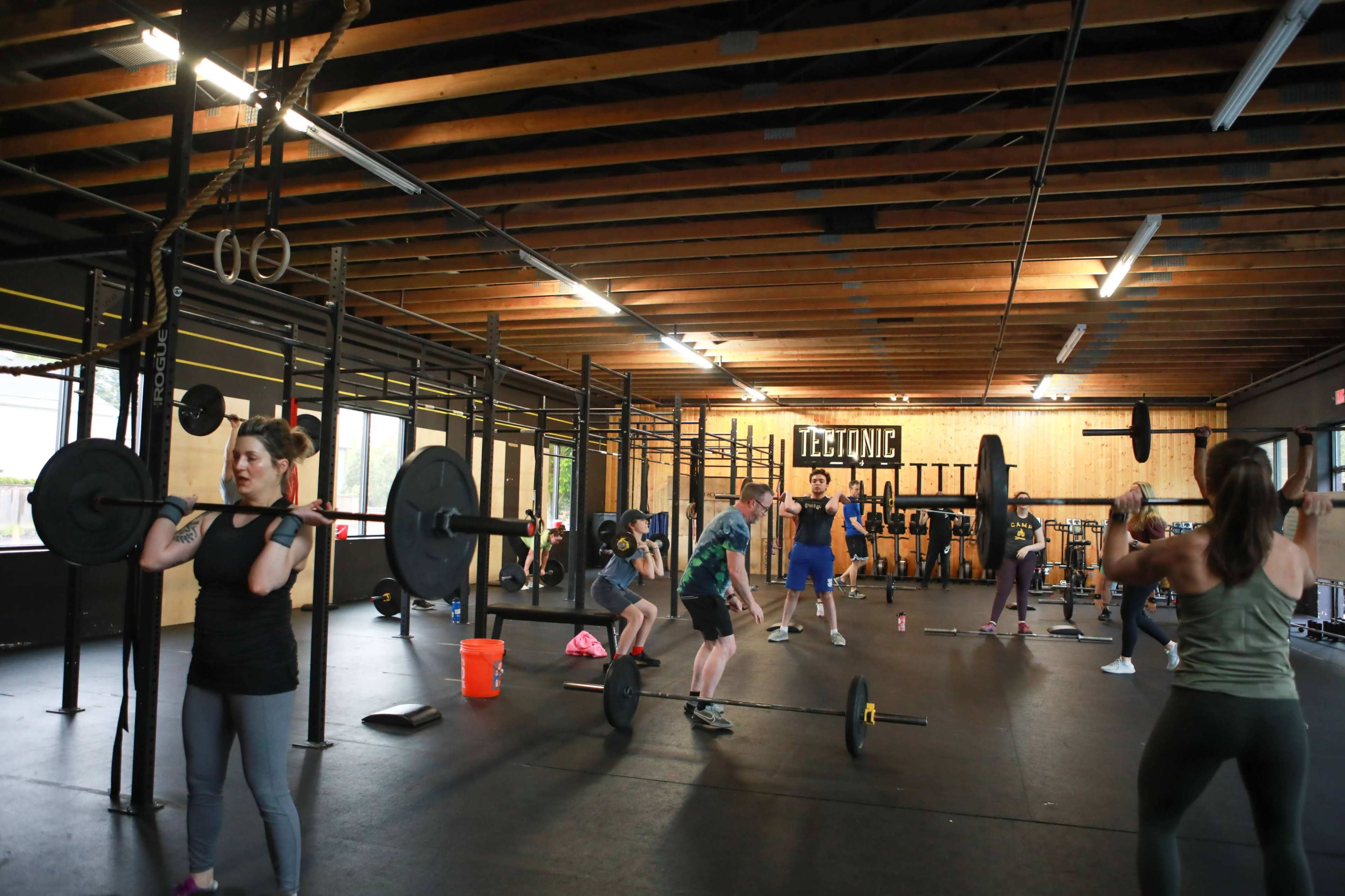 A group of people is engaged in various weightlifting exercises in a gym with a wooden ceiling and exercise equipment in the background.