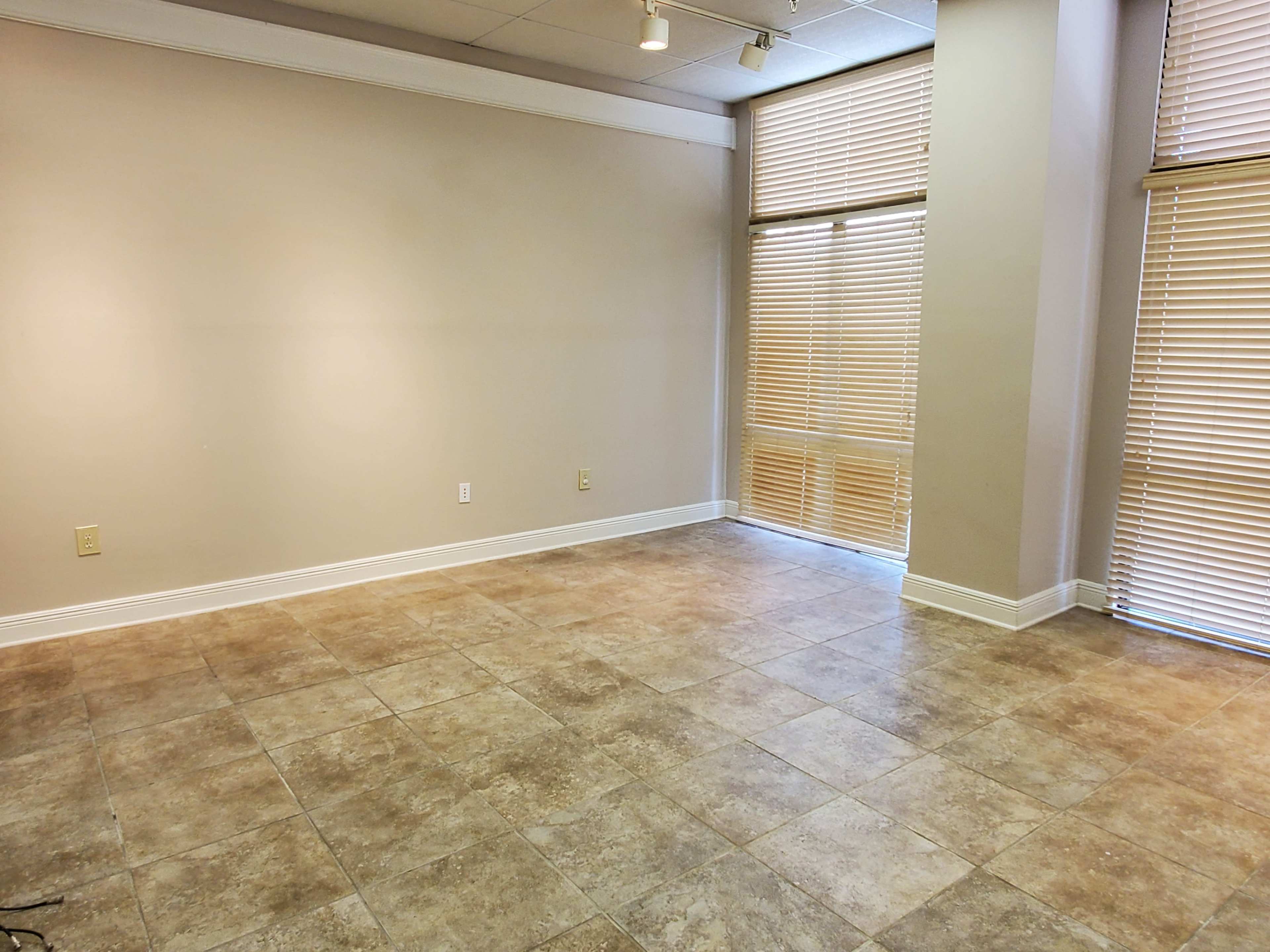 The image shows an empty room with tiled flooring and large windows covered by blinds.