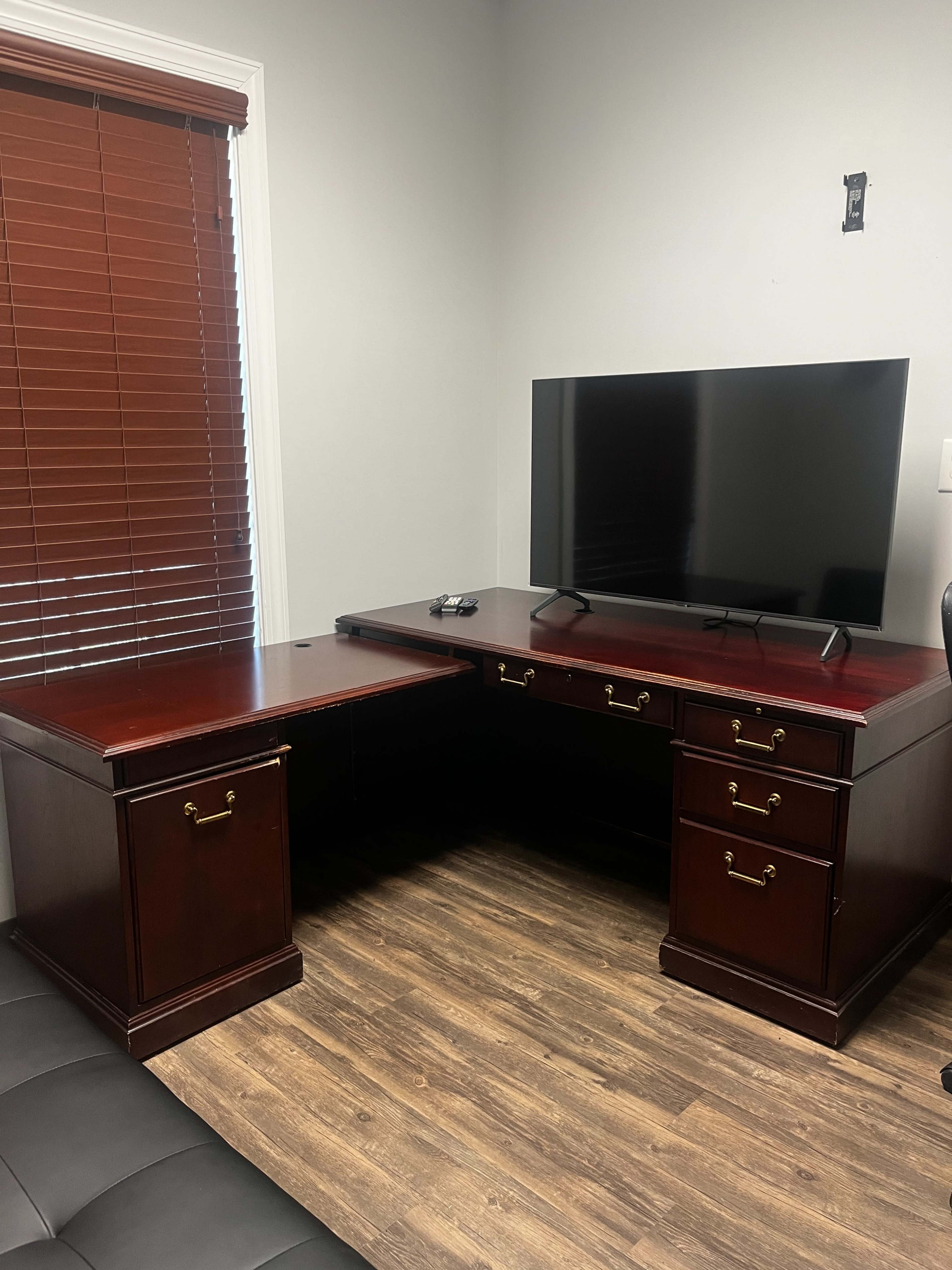 The image shows a corner office setup featuring a dark wooden desk with drawers and a television mounted on the wall above it.