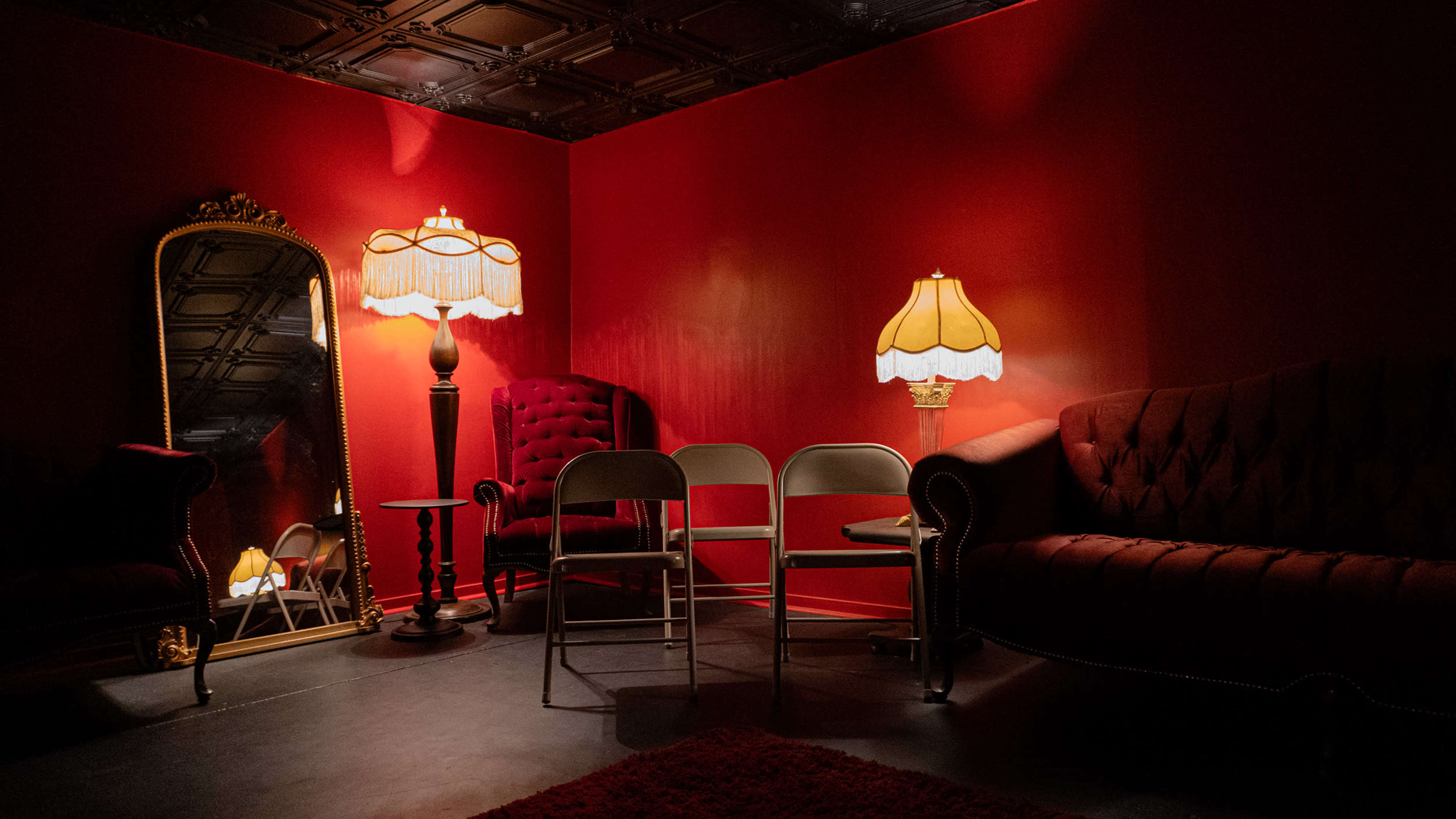 A dimly lit room features red walls, vintage furnishings, a large mirror, and two illuminated lamps.