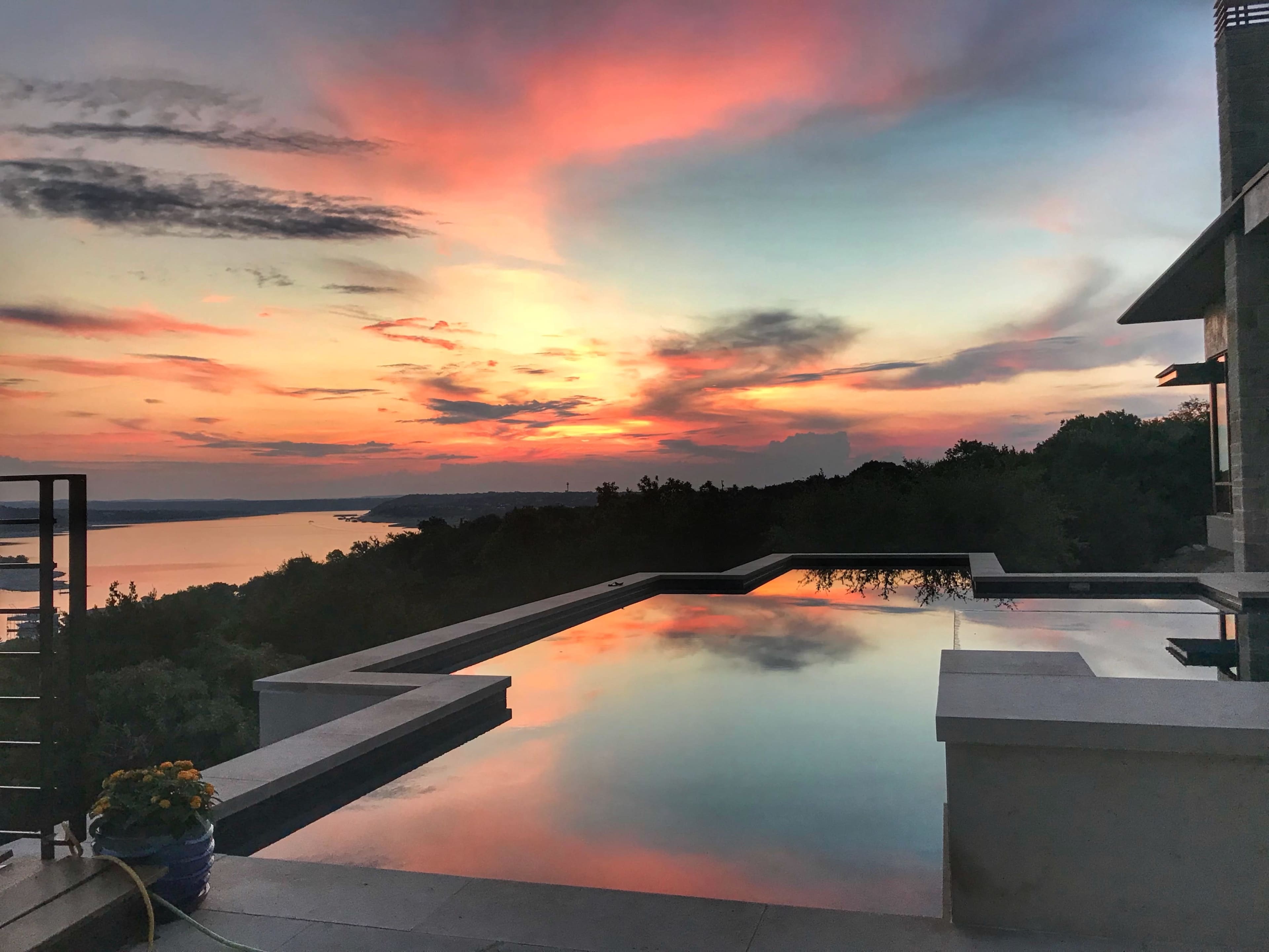 The image shows a tranquil infinity pool reflecting a colorful sunset over a lake and surrounding landscape.