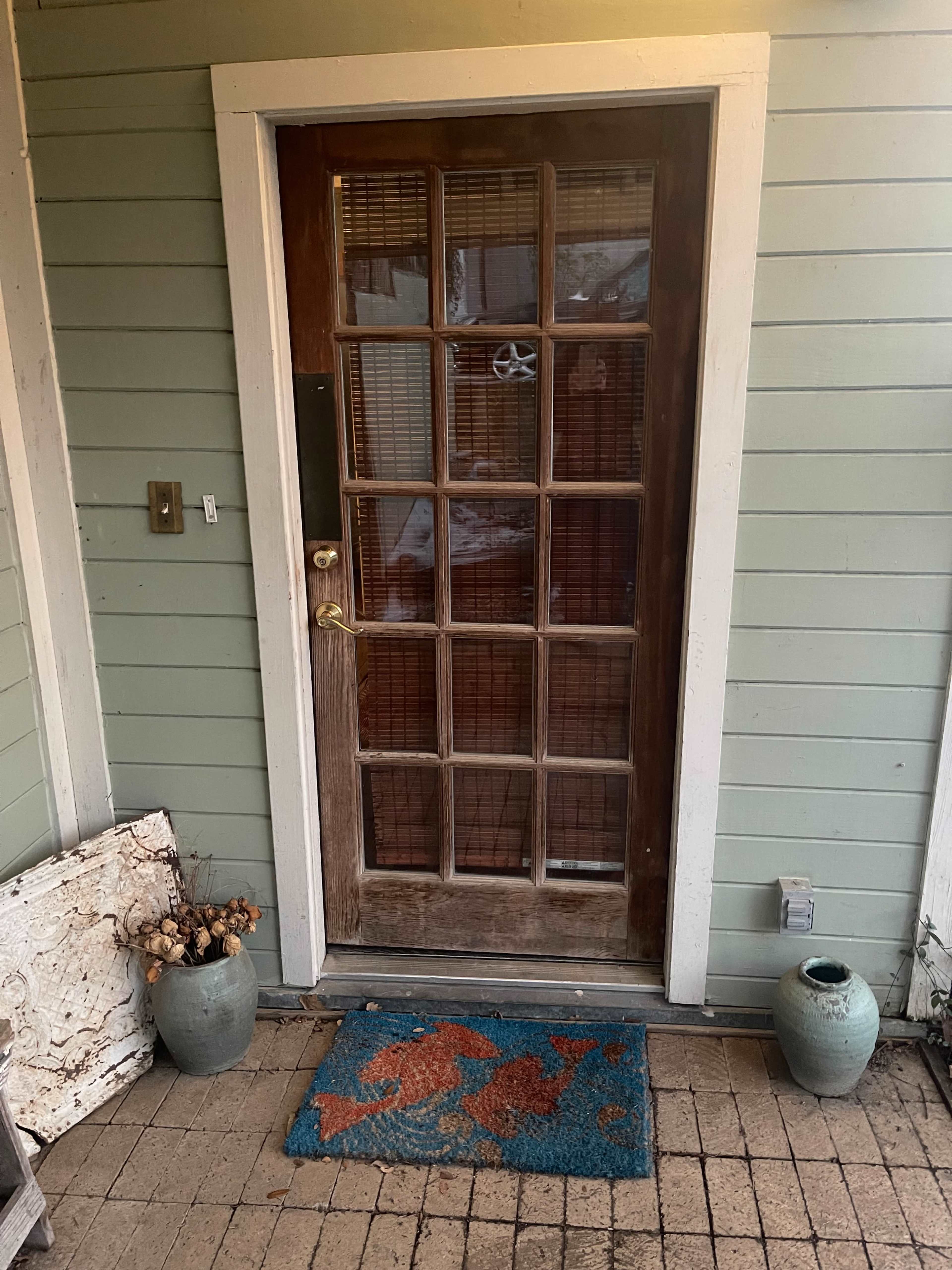 A wooden door with multiple glass panes, framed by green siding, and a welcome mat on a brick walkway in front.