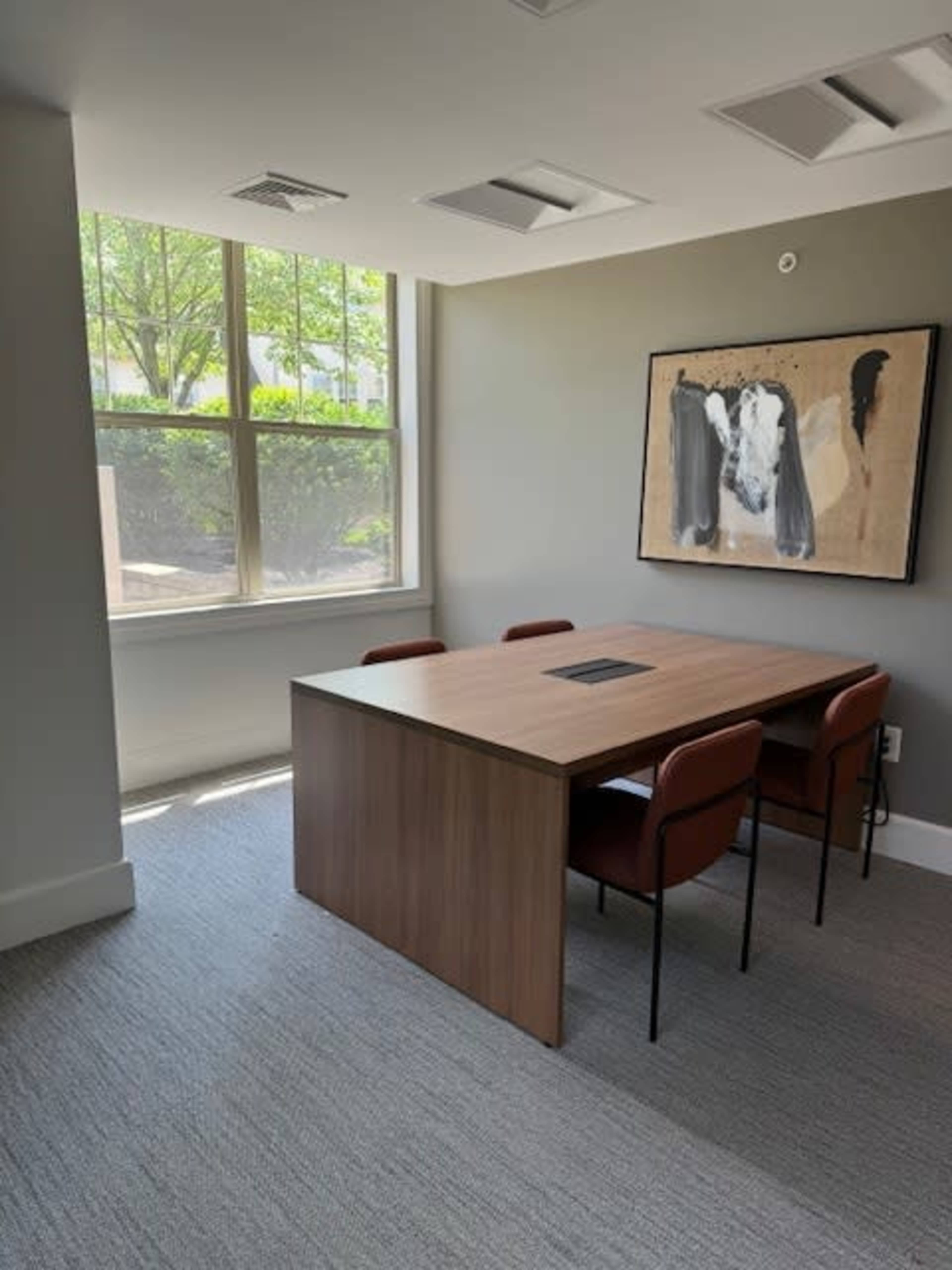 A small conference room featuring a wooden table surrounded by four chairs, with large windows letting in natural light and a piece of art on the wall.